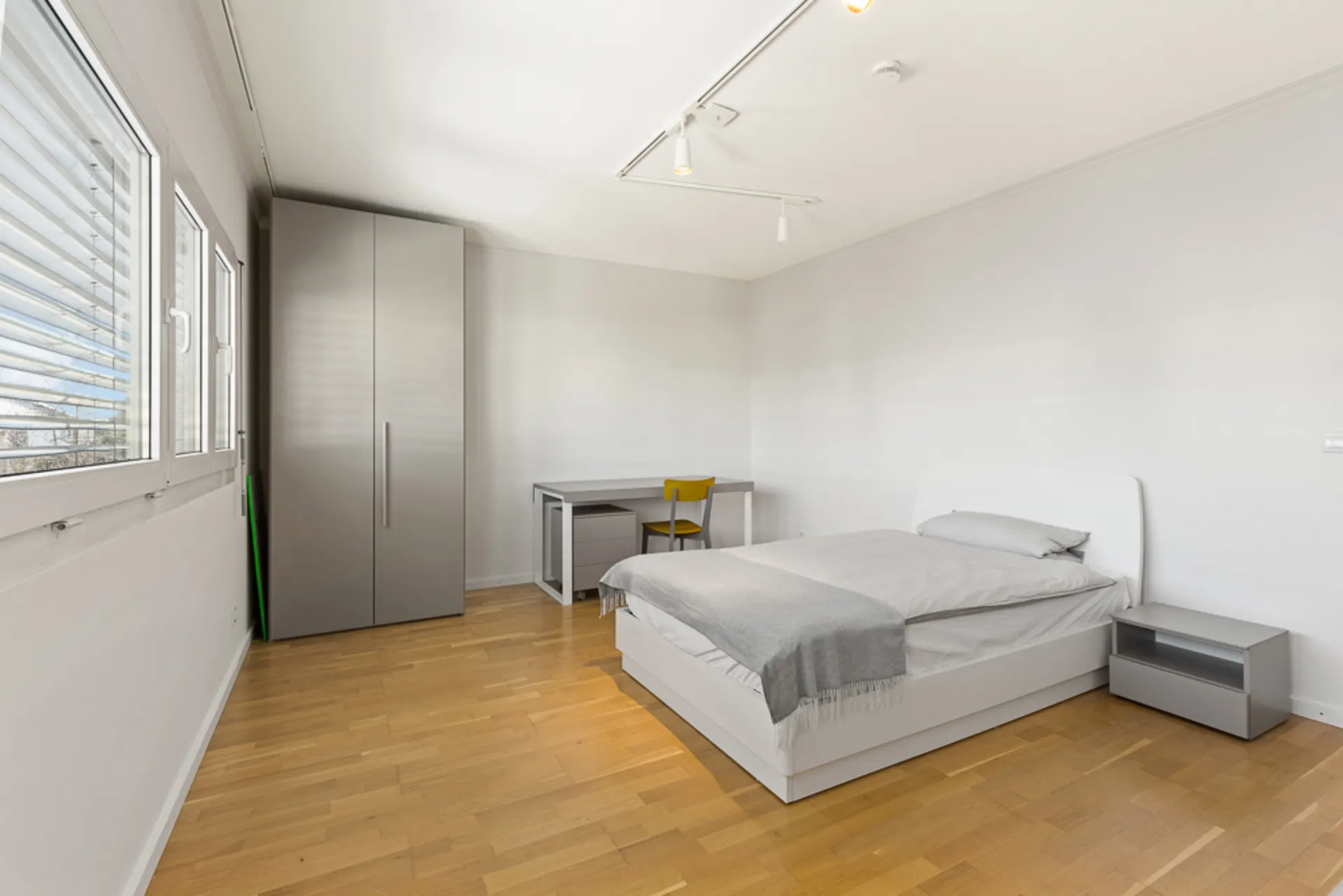 Modernes Schlafzimmer mit Doppelbett, grauem Kleiderschrank, Schreibtisch und Nachttisch auf Parkettboden.