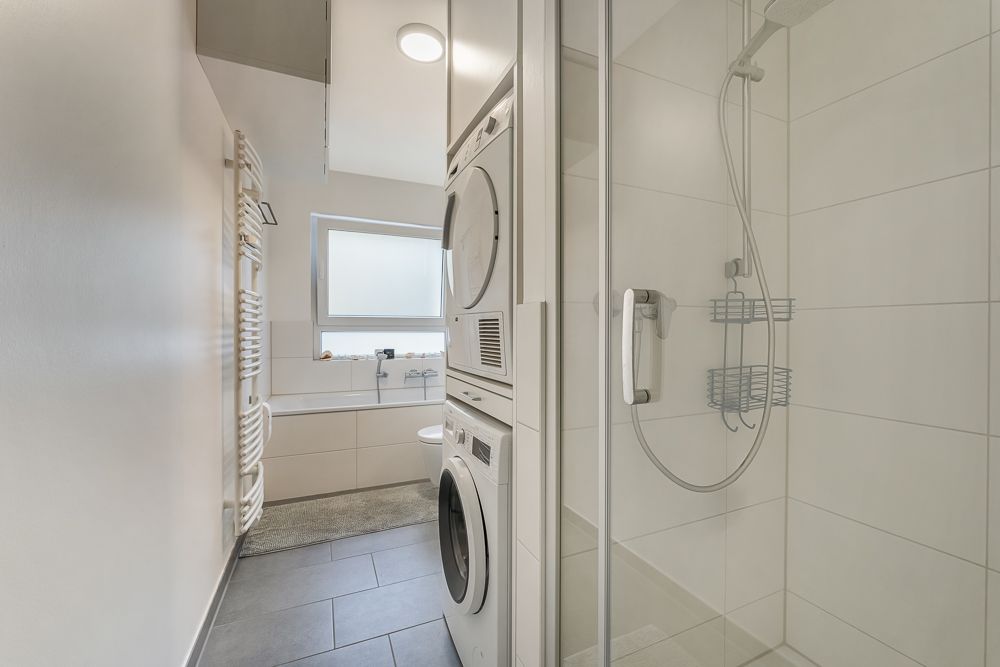 Modernes Badezimmer mit Dusche, Waschmaschine und Trockner, Fenster und Heizkörper.