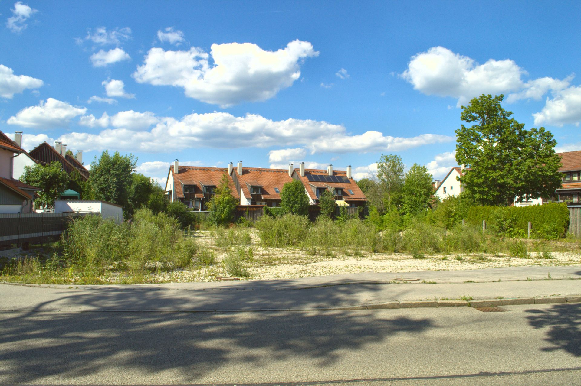 Leeres Grundstück mit etwas Bewuchs vor Wohnhäusern mit roten Dächern bei blauem Himmel.