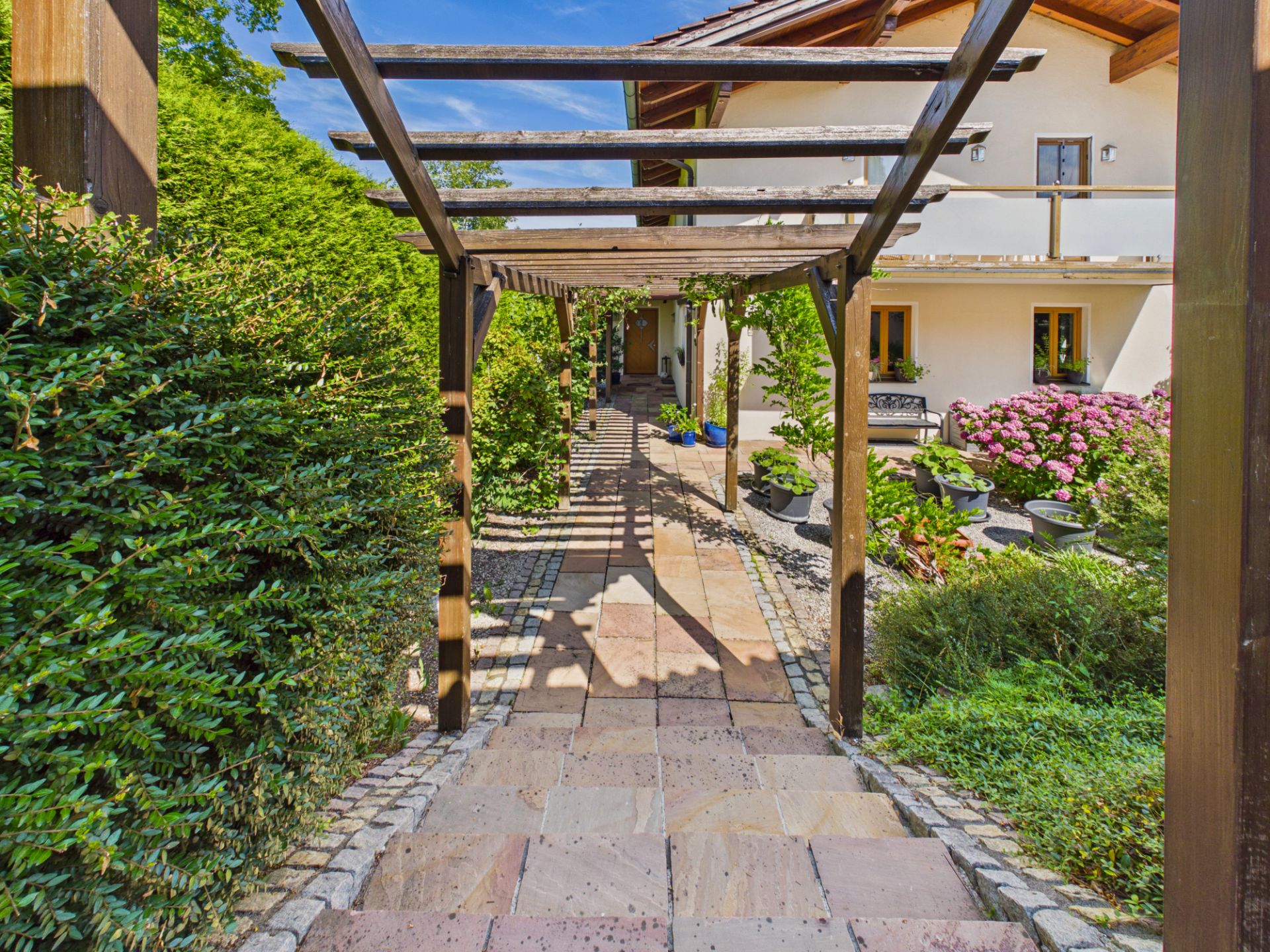Gepflasterter Weg mit Pergola, umgeben von Büschen und Blumen, führt zum Hauseingang.