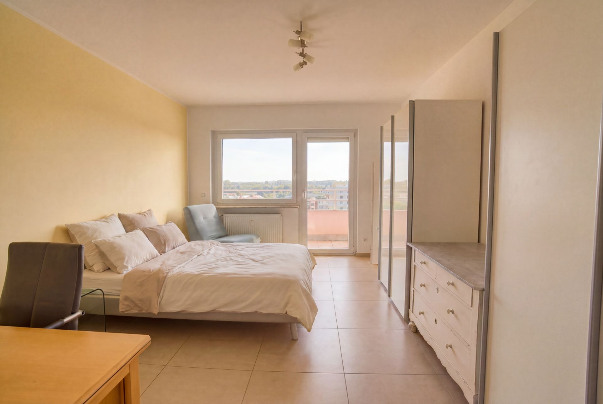 Helles Schlafzimmer mit Bett, Schreibtisch, Schrank, Kommode und großem Fenster mit Balkon.