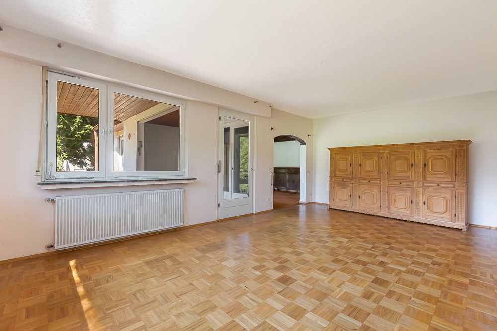 Helles Wohnzimmer mit Parkettboden, großem Fenster, Glastür und Holzschrank an der Wand.