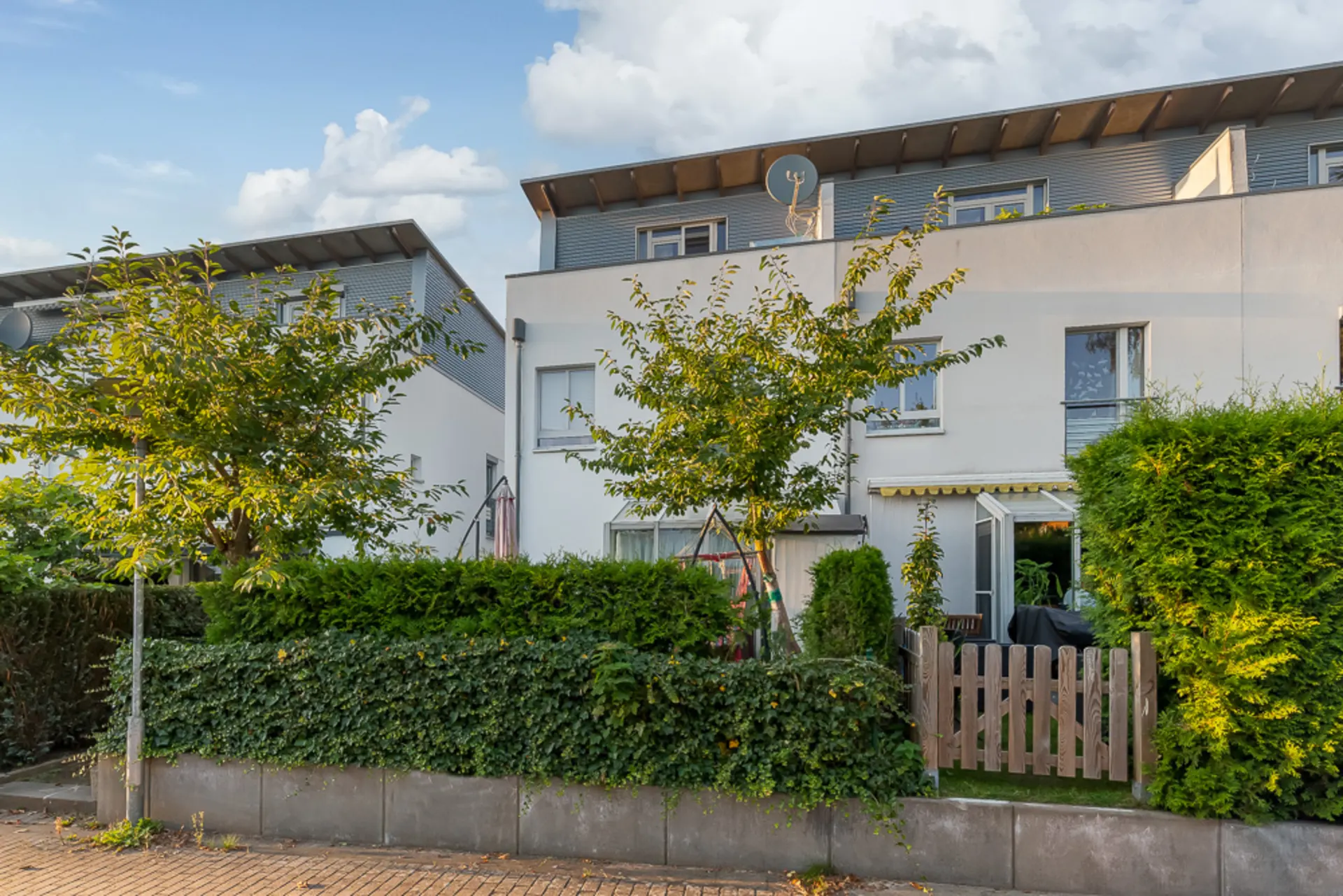 Moderne Reihenhausfassade mit Garten, Bäumen und kleinem Holzzaun im Vordergrund.