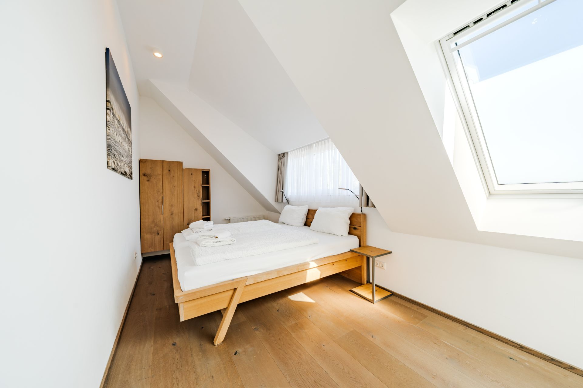 Helles Schlafzimmer mit Doppelbett, Holzschrank und großem Fenster auf Holzfußboden.