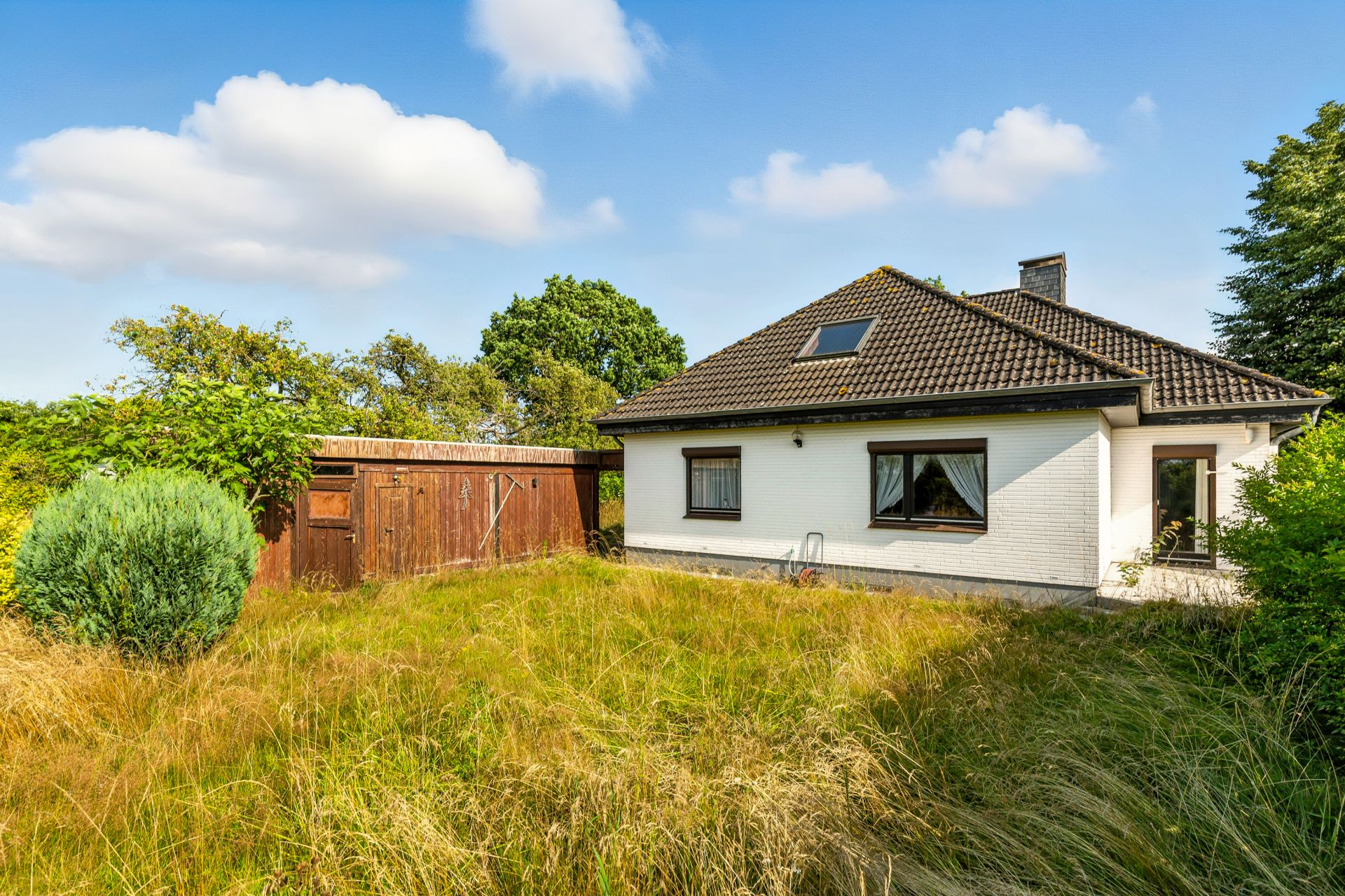 Weißes Einfamilienhaus mit dunklem Dach und großem Garten mit hohem Gras, Holzschuppen links.