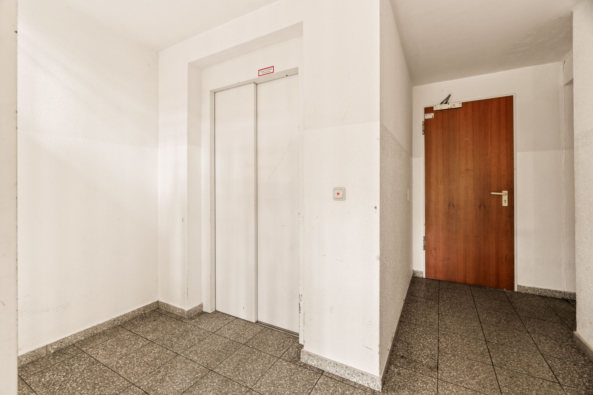 Heller Flur mit Aufzugstür und brauner Wohnungstür, Fliesenboden.