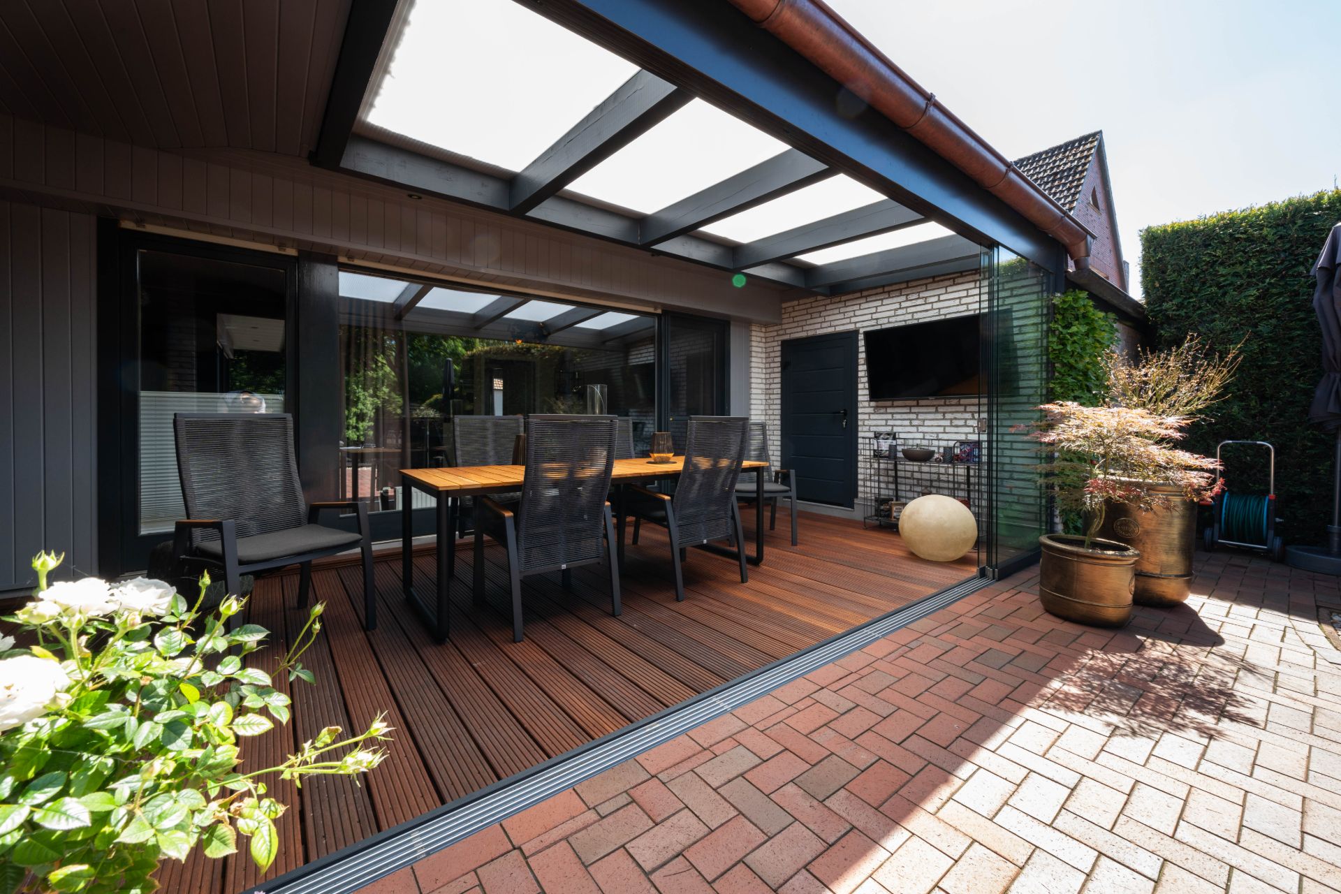 Überdachte Terrasse mit Holzboden, Tisch, Stühlen und Pflanzen neben einem Haus.