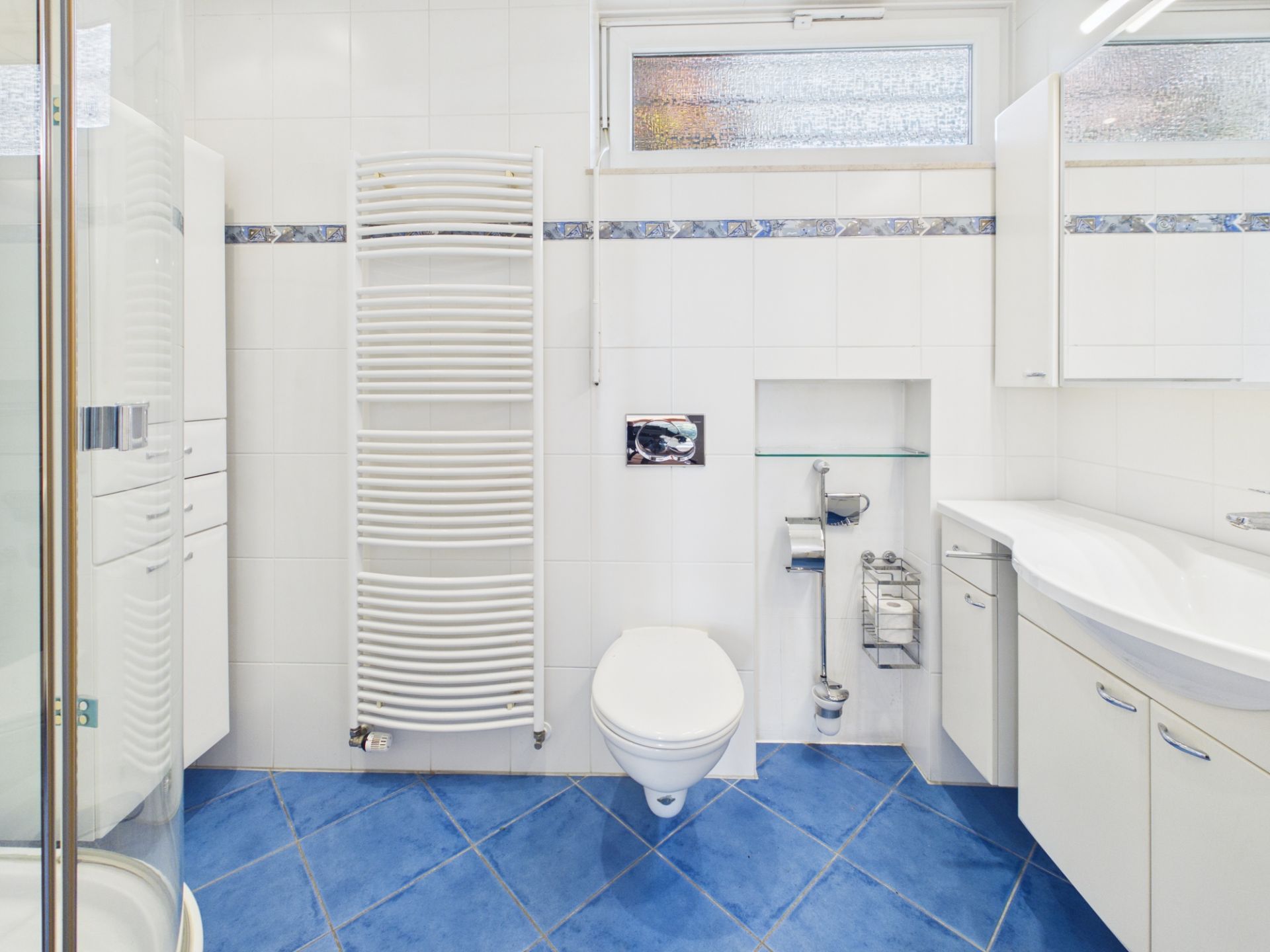 Modernes Bad mit blauem Fliesenboden, weißer Wand, Hängetoilette und Handtuchheizkörper.