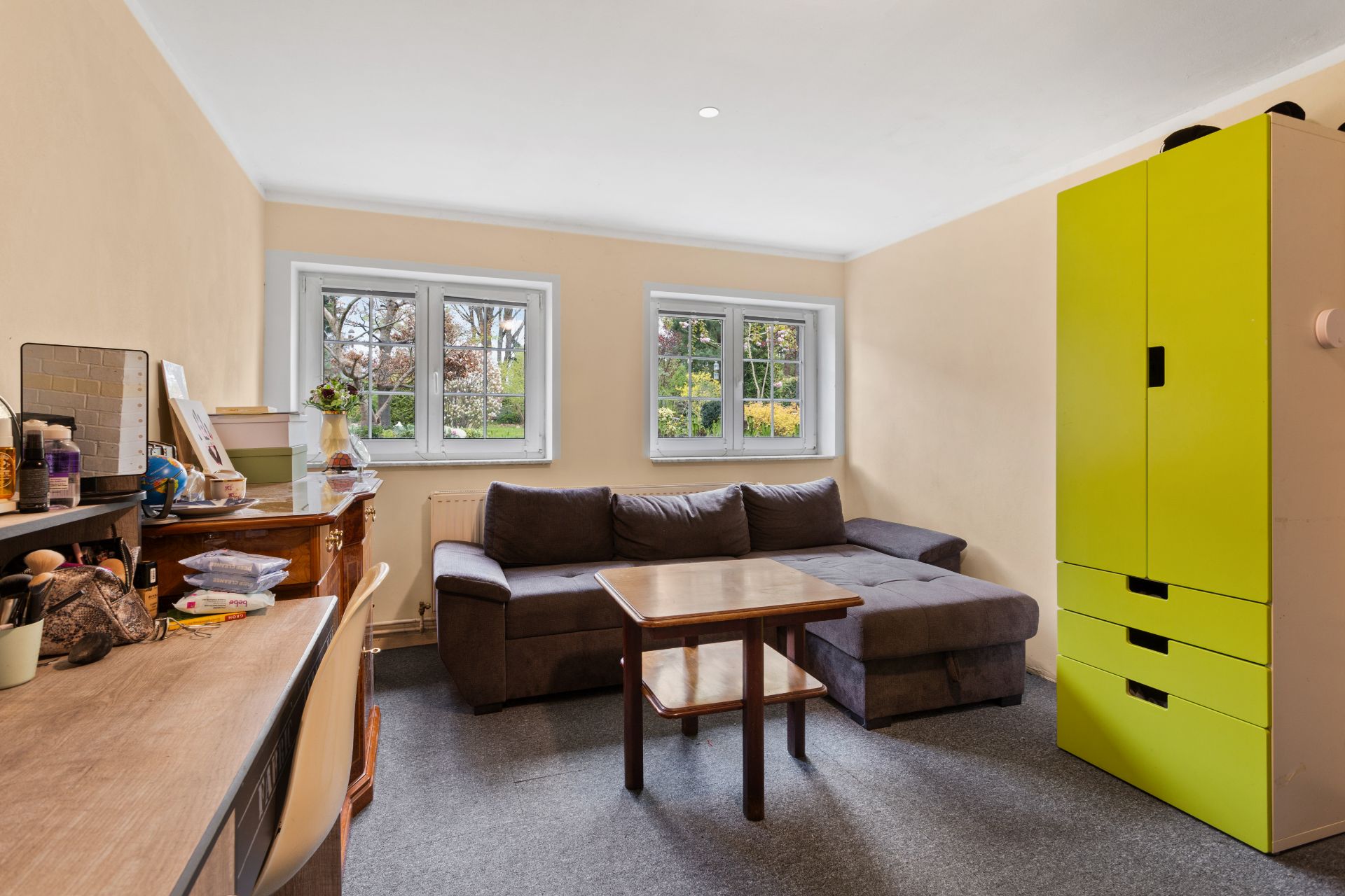 Wohnzimmer mit grauem Sofa, Holztisch, Schreibtisch und grünem Schrank, drei Fenster im Hintergrund.