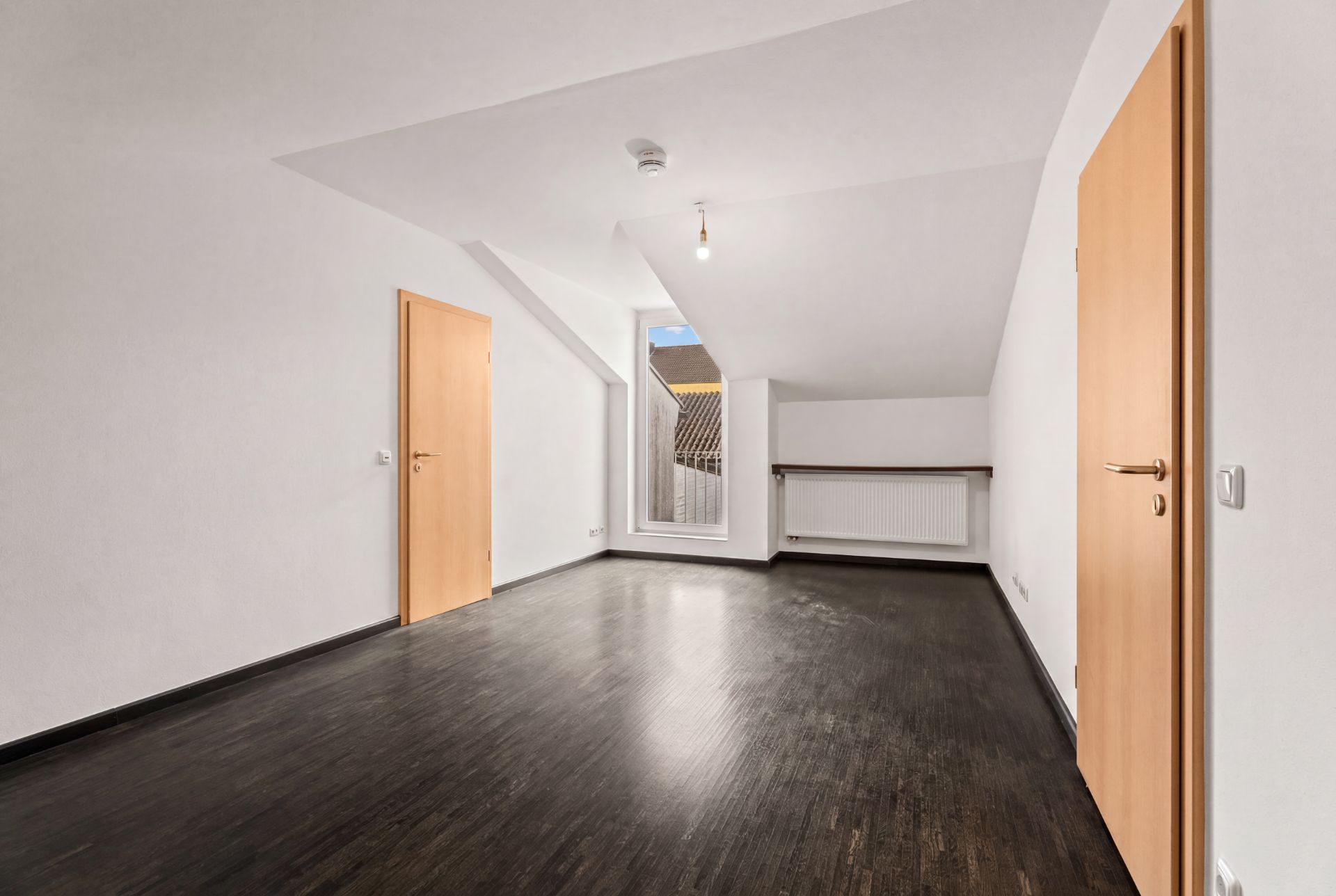 Leerer Raum mit dunklem Boden, weißen Wänden, zwei Holztüren und einem Fenster unter einer Schräge.