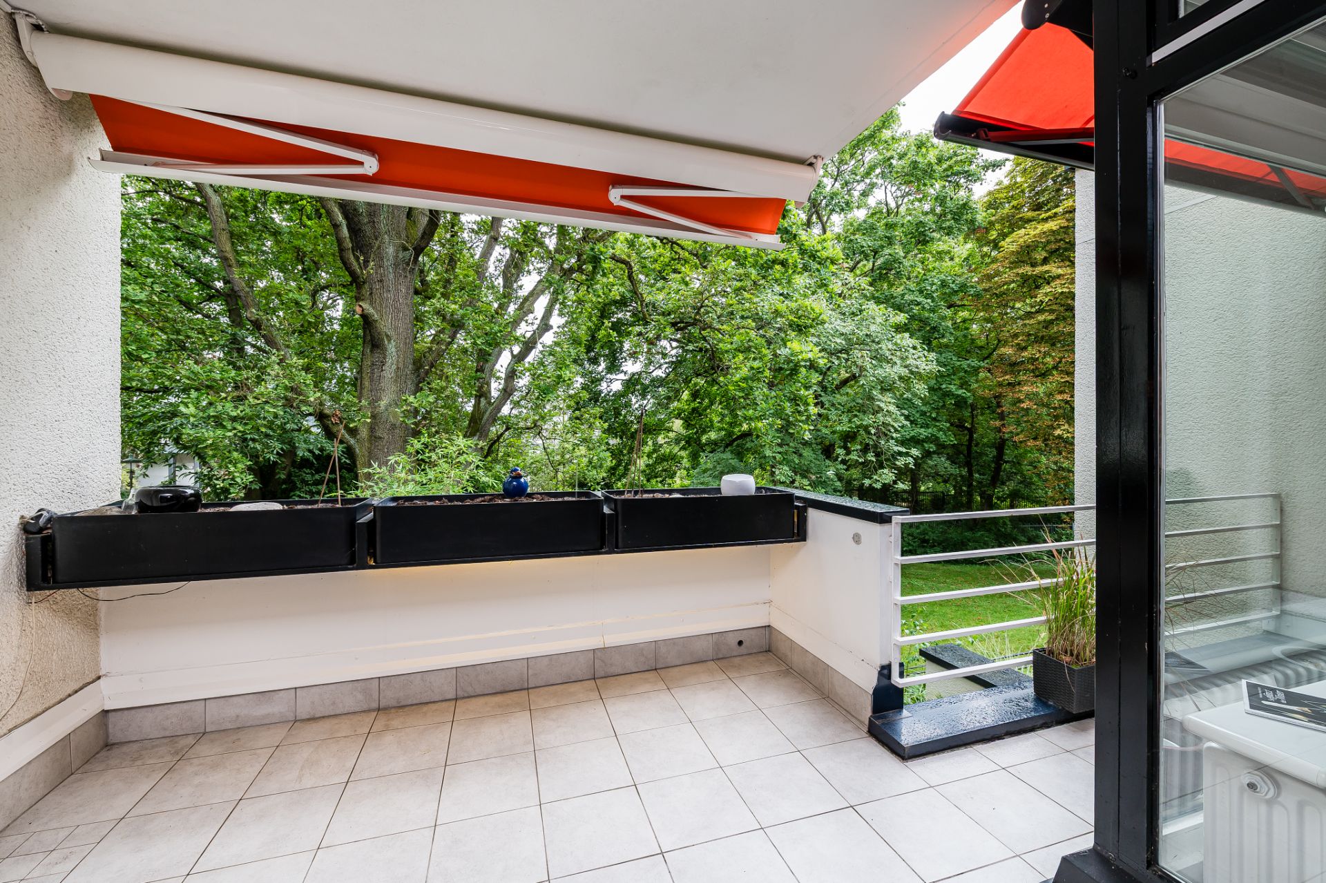 Großer Balkon mit hellen Fliesen, Markise und Blick auf grüne Bäume im Garten.
