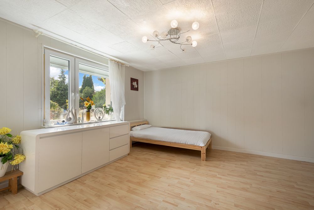 Helles Schlafzimmer mit Holzboden, Bett, großem Fenster und Kommode, dekoriert mit Blumen.