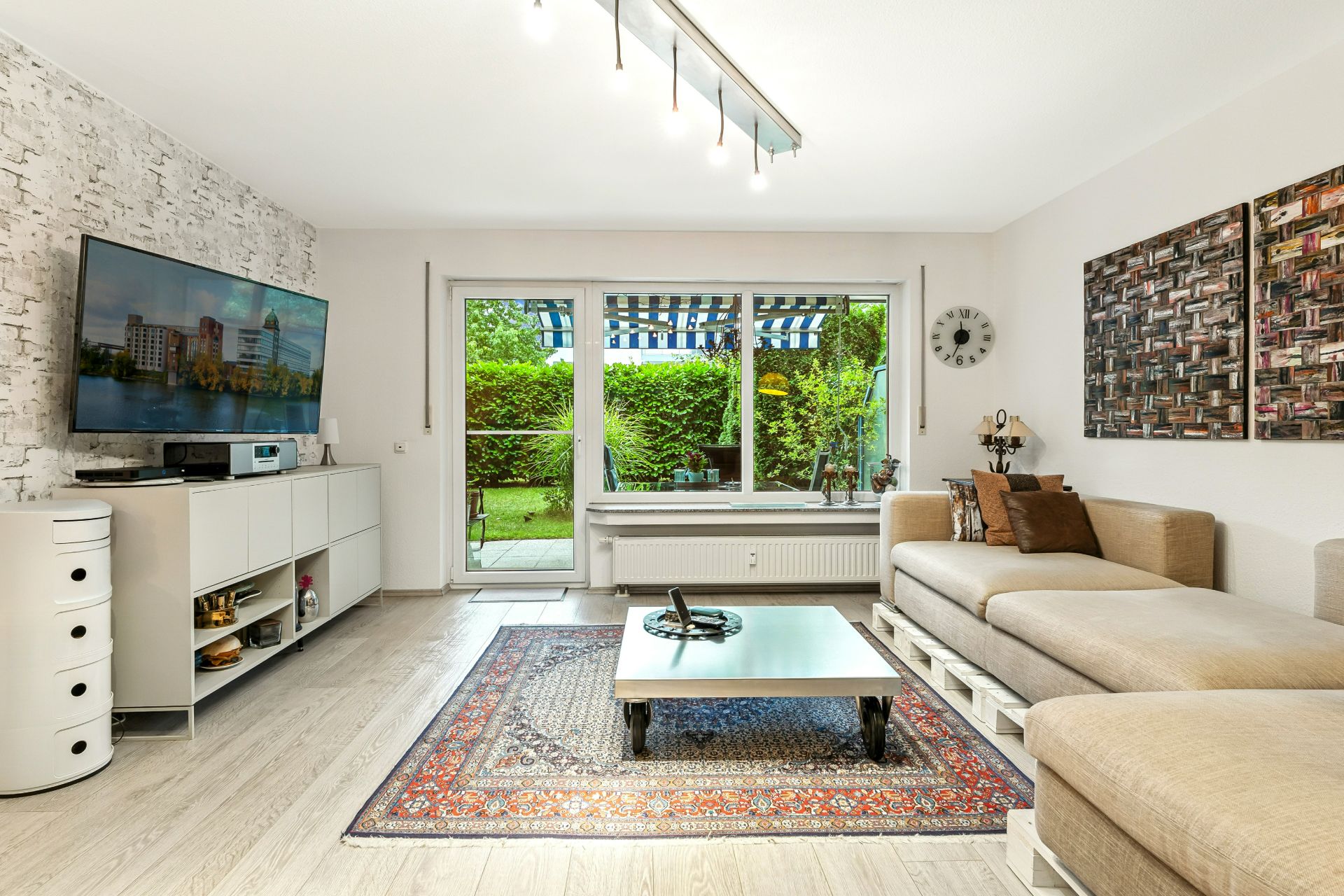 Helles Wohnzimmer mit Sofa, Couchtisch, Teppich und Blick auf Garten durch große Fenster.