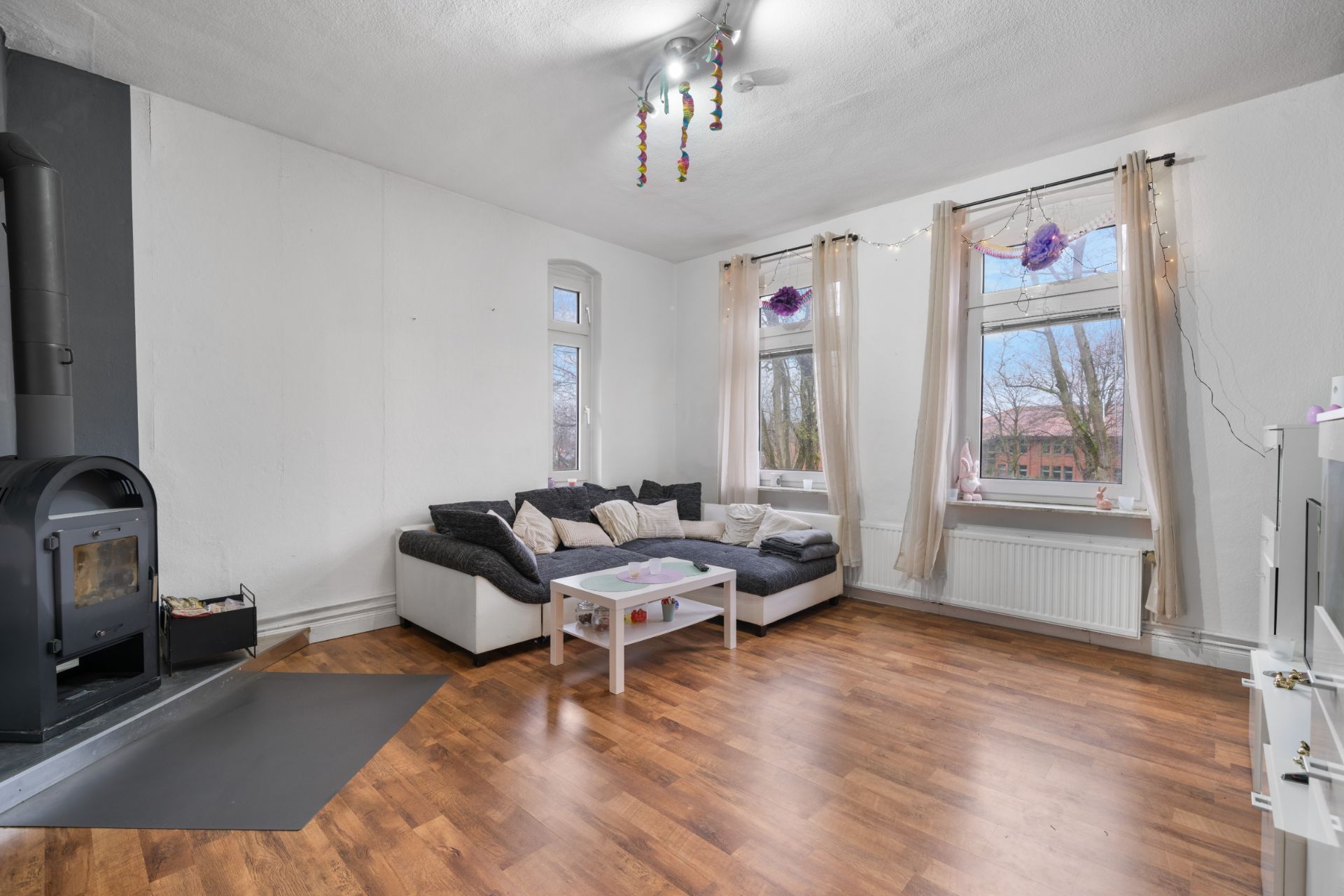 Helles Wohnzimmer mit Ecksofa, Couchtisch, Holzofen und großen Fenstern mit Vorhängen.