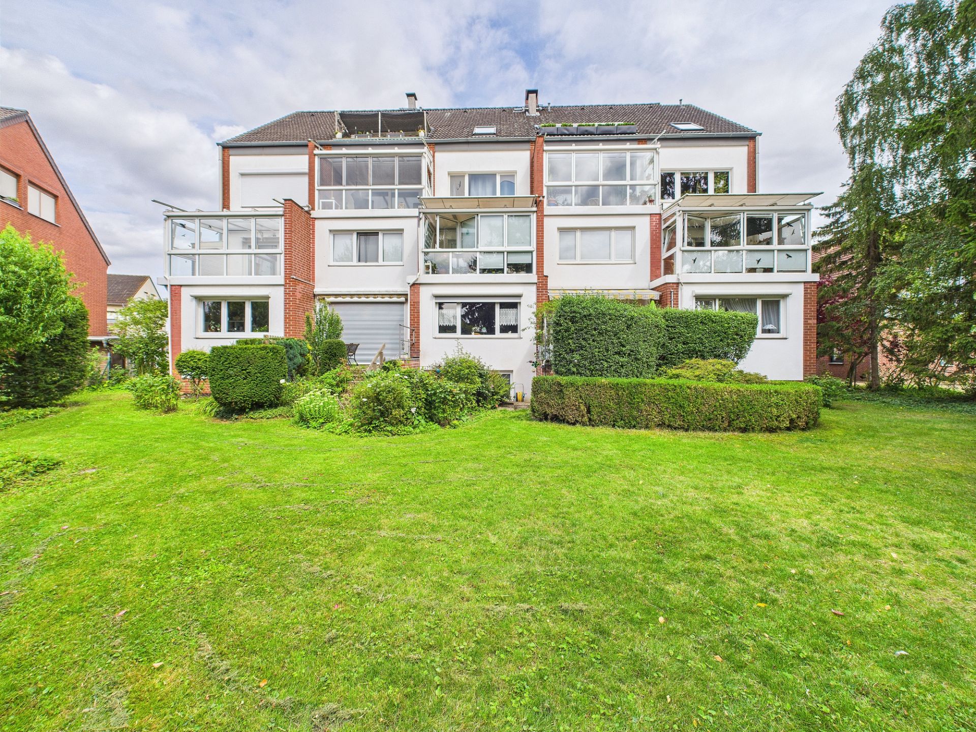 Mehrfamilienhaus mit großen Fenstern und Wintergärten, davor gepflegter Garten mit Büschen und Rasen.