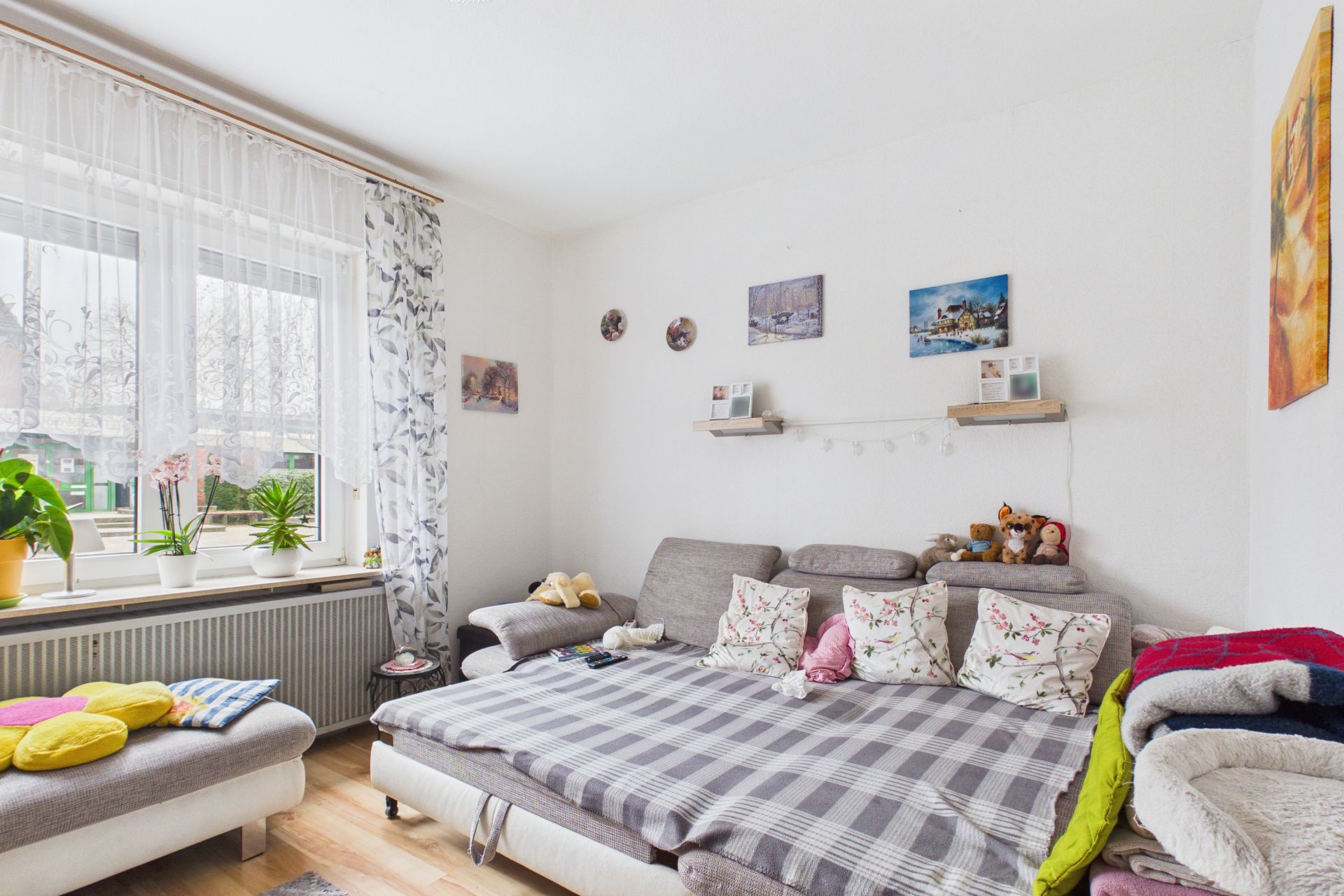 Helles Wohnzimmer mit Sofa, vielen Kissen, Pflanzen am Fenster und Wanddekoration.