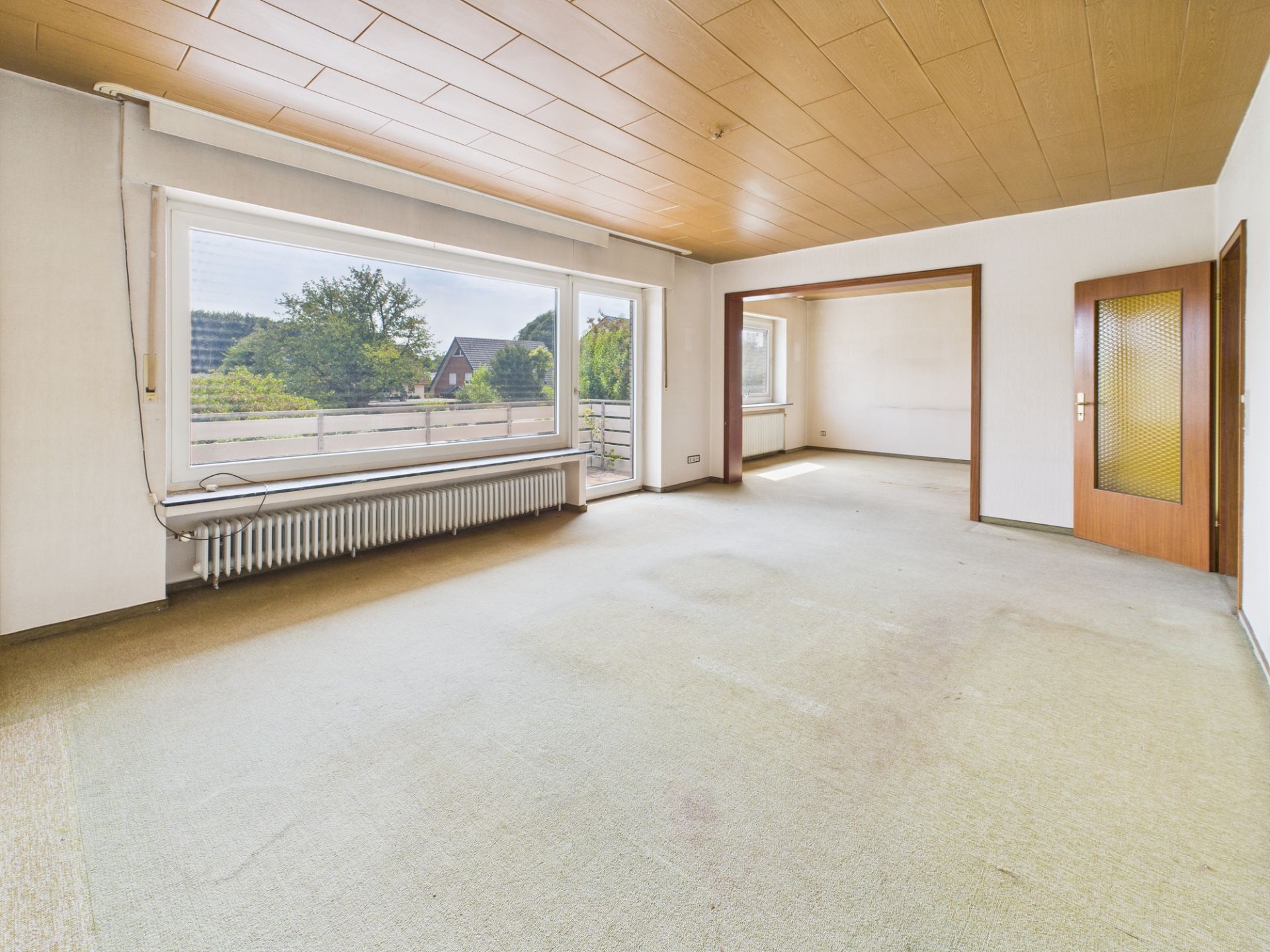 Leeres Wohnzimmer mit großen Fenstern, Blick auf Garten, Holzdecke und Tür zu weiterem Raum.