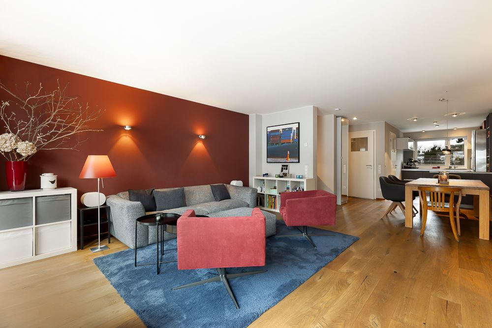 Helles Wohnzimmer mit roter Wand, grauem Sofa, zwei roten Sesseln und Essbereich im Hintergrund.