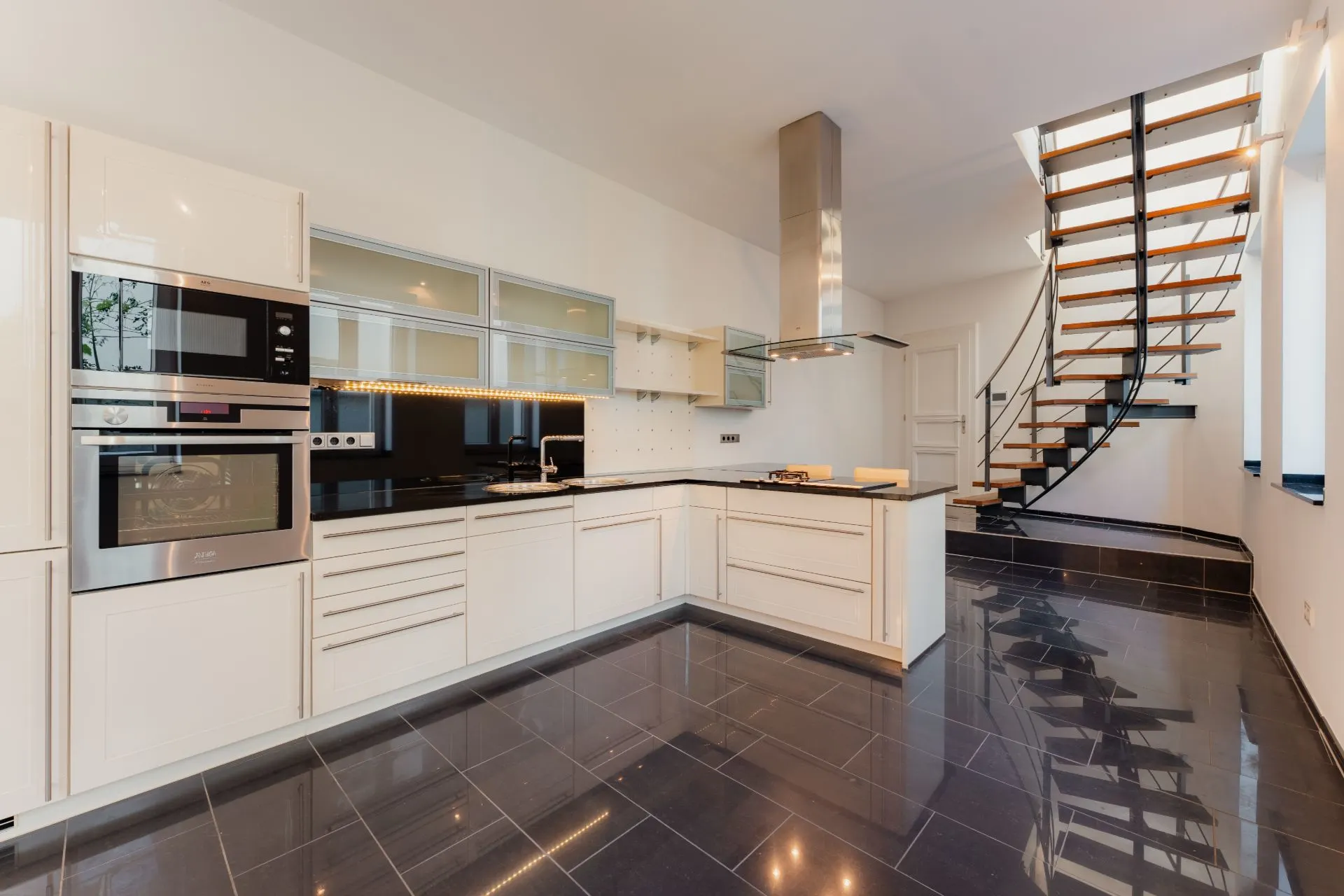 Moderne Einbauküche mit schwarzen Fliesen und offener Holztreppe im Hintergrund.