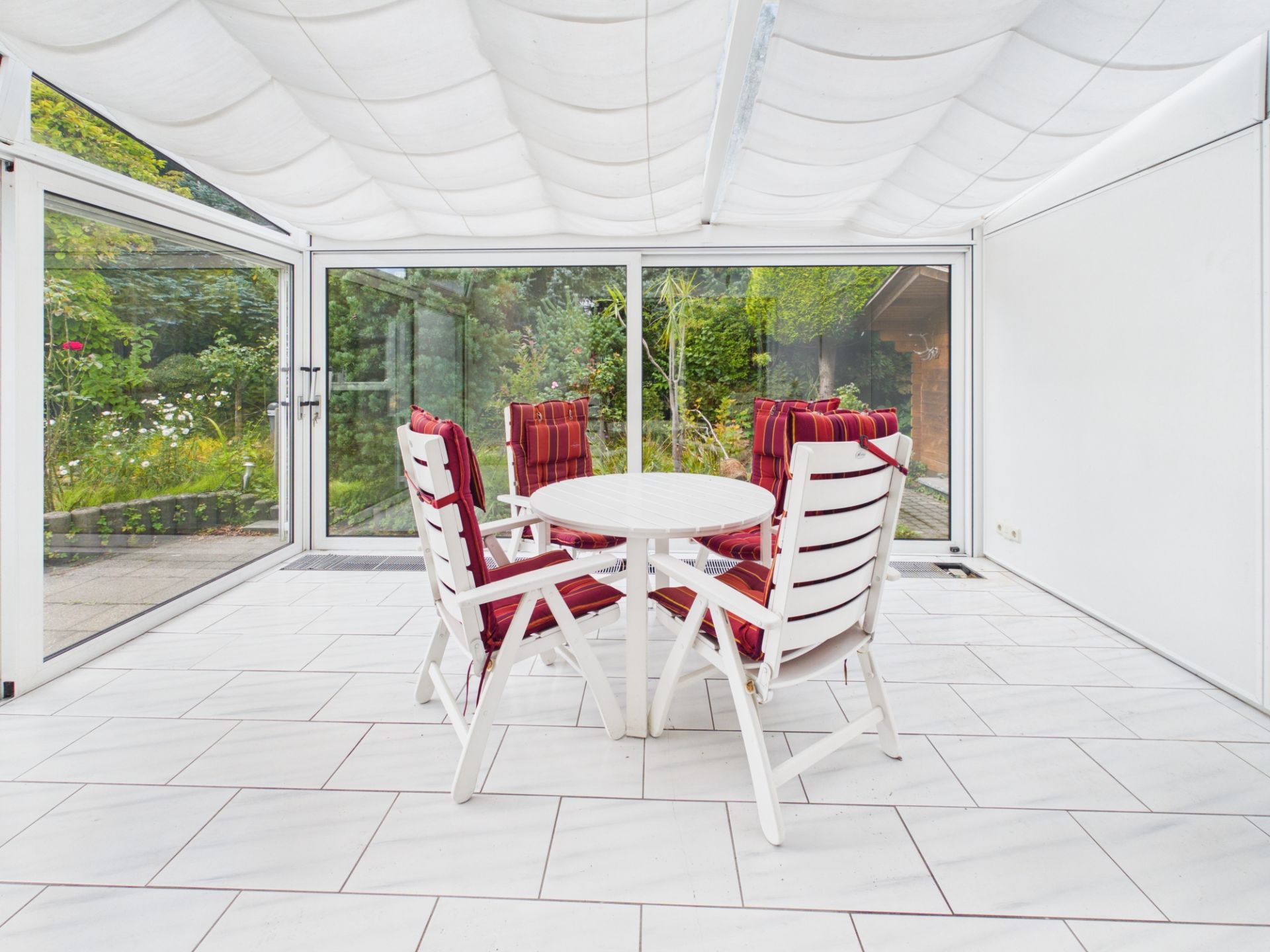 Heller Wintergarten mit weißen Stühlen und rundem Tisch, Blick in einen Garten.