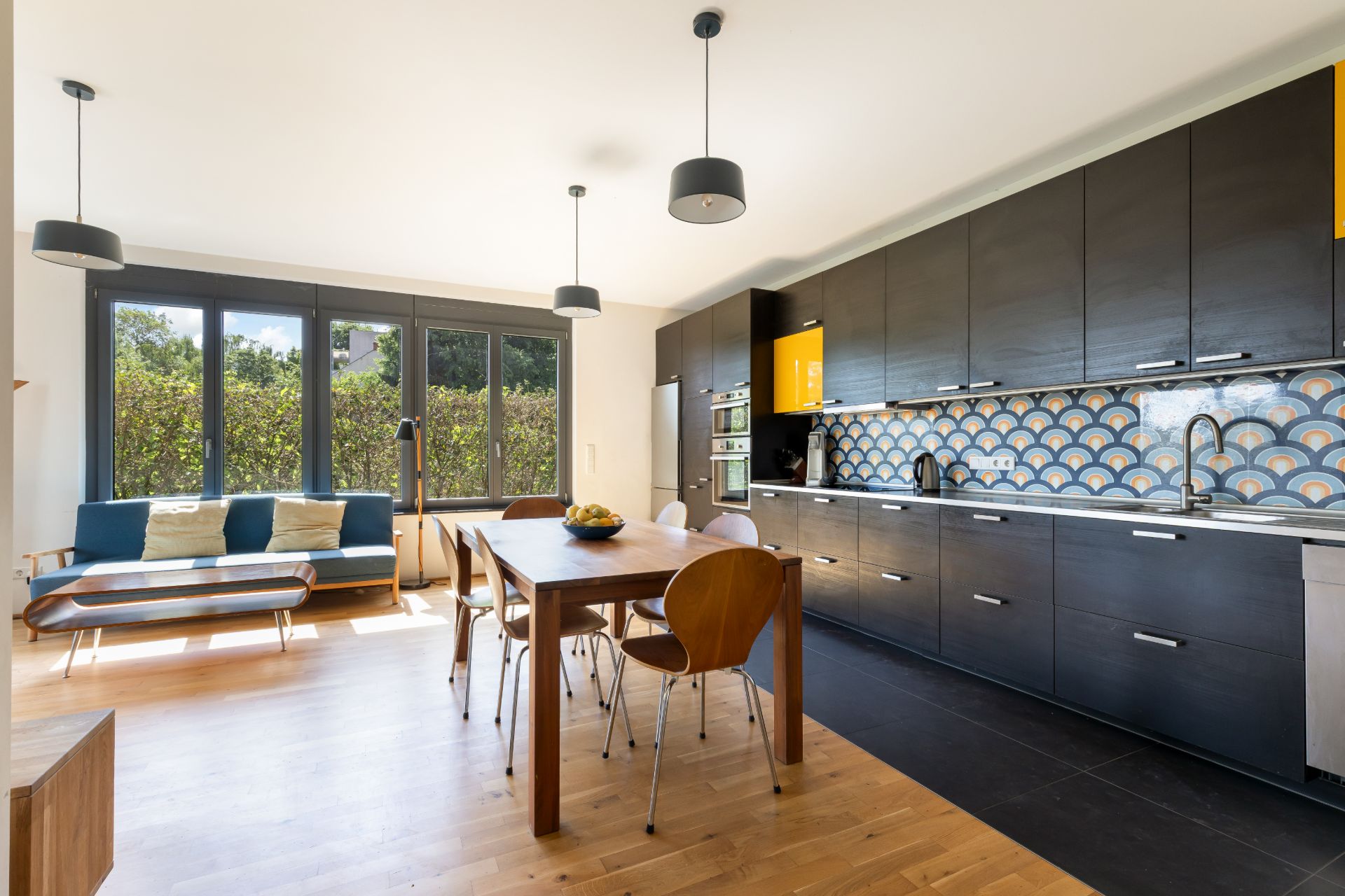 Moderne Küche mit dunklen Schränken, Holztisch, Stühlen und Sofa vor großen Fenstern.
