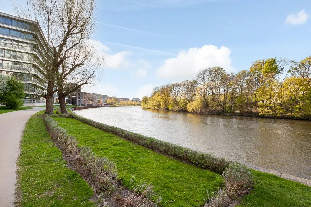 Grüner Weg und Fluss mit Bäumen, daneben modernes Wohngebäude.