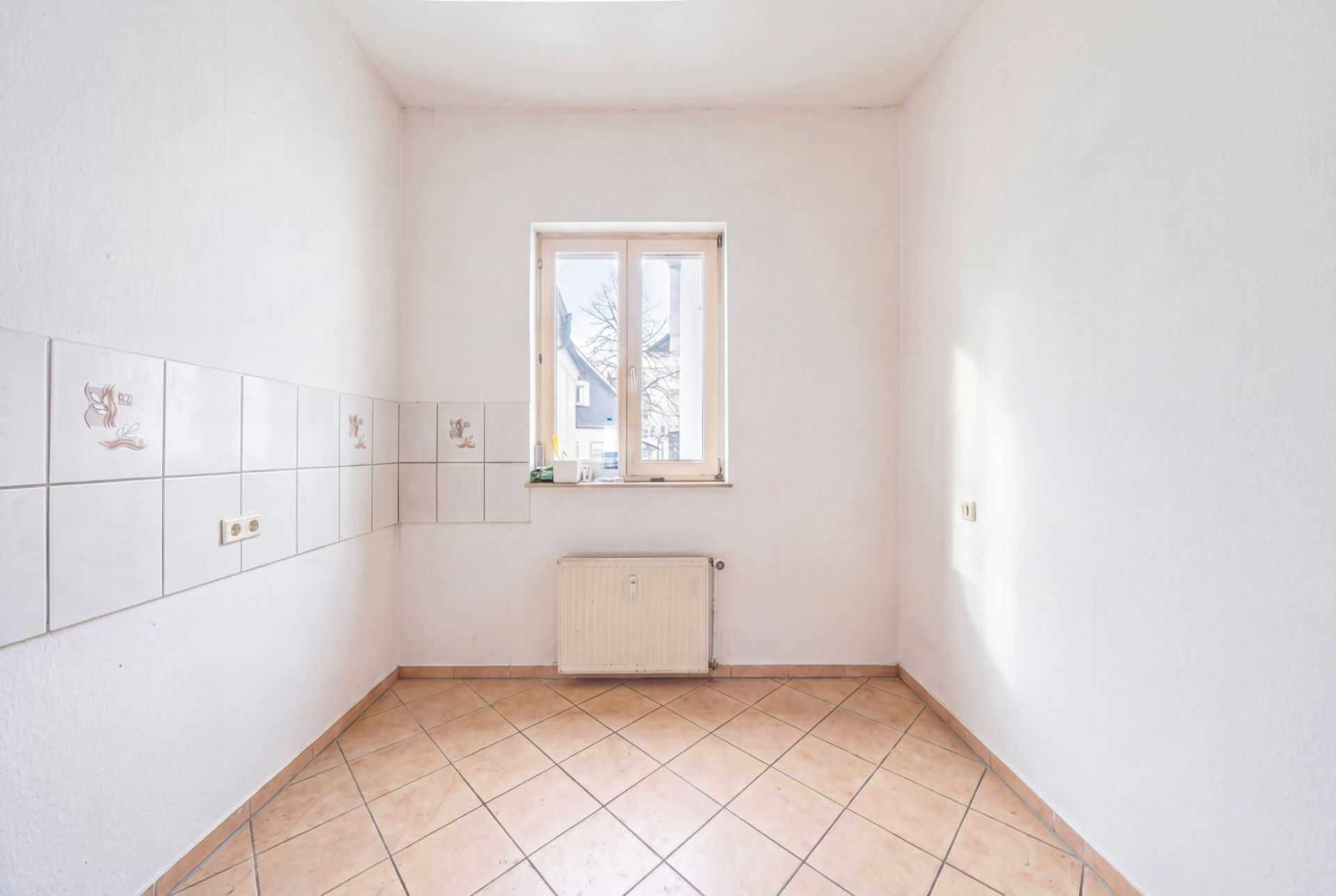 Leerer, heller Raum mit Fliesenboden, weißen Wänden und Fenster, Wandfliesen auf der linken Seite.