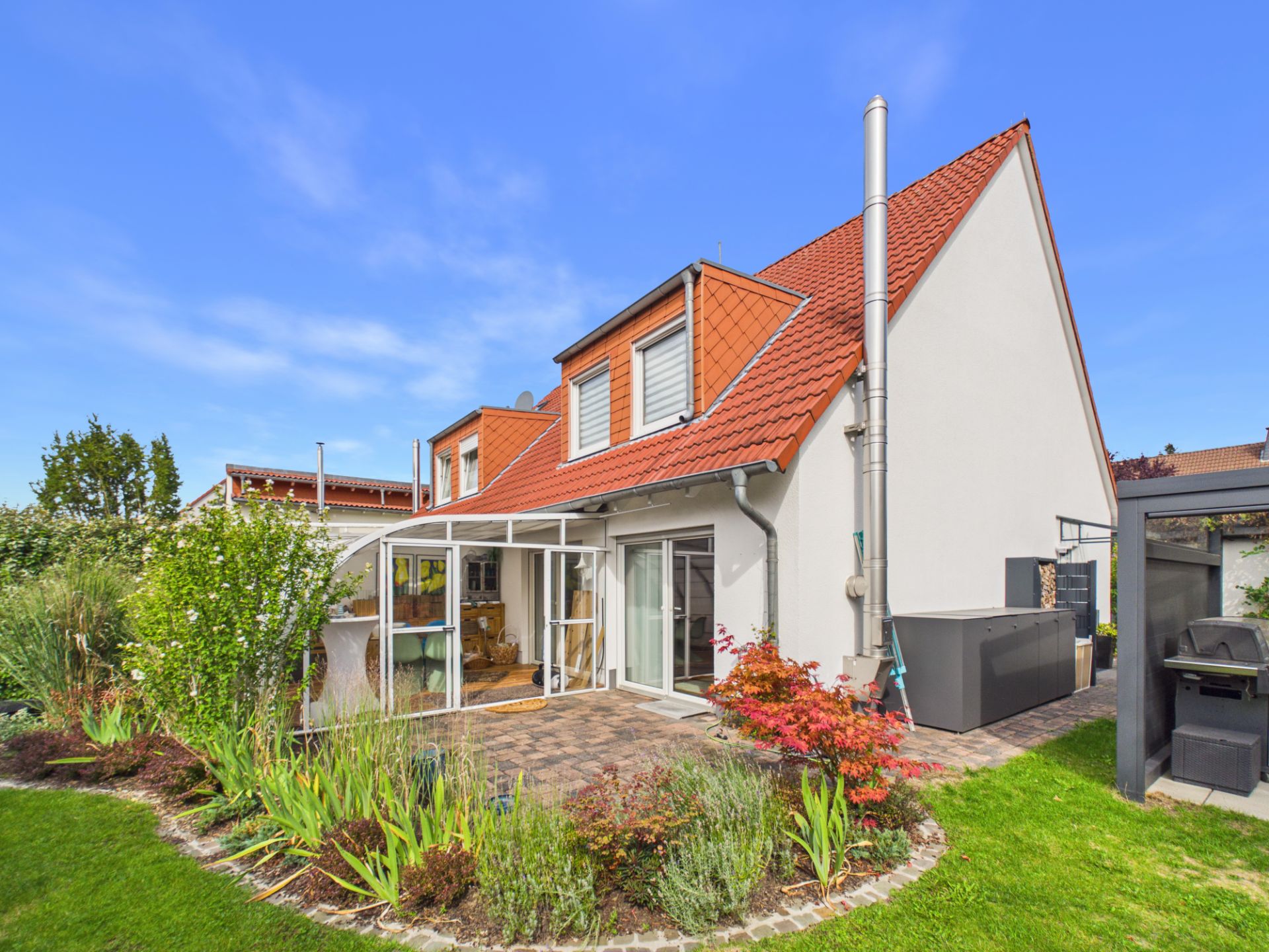 Helles Einfamilienhaus mit Wintergarten, rotem Dach und gepflegtem Garten im Vordergrund.
