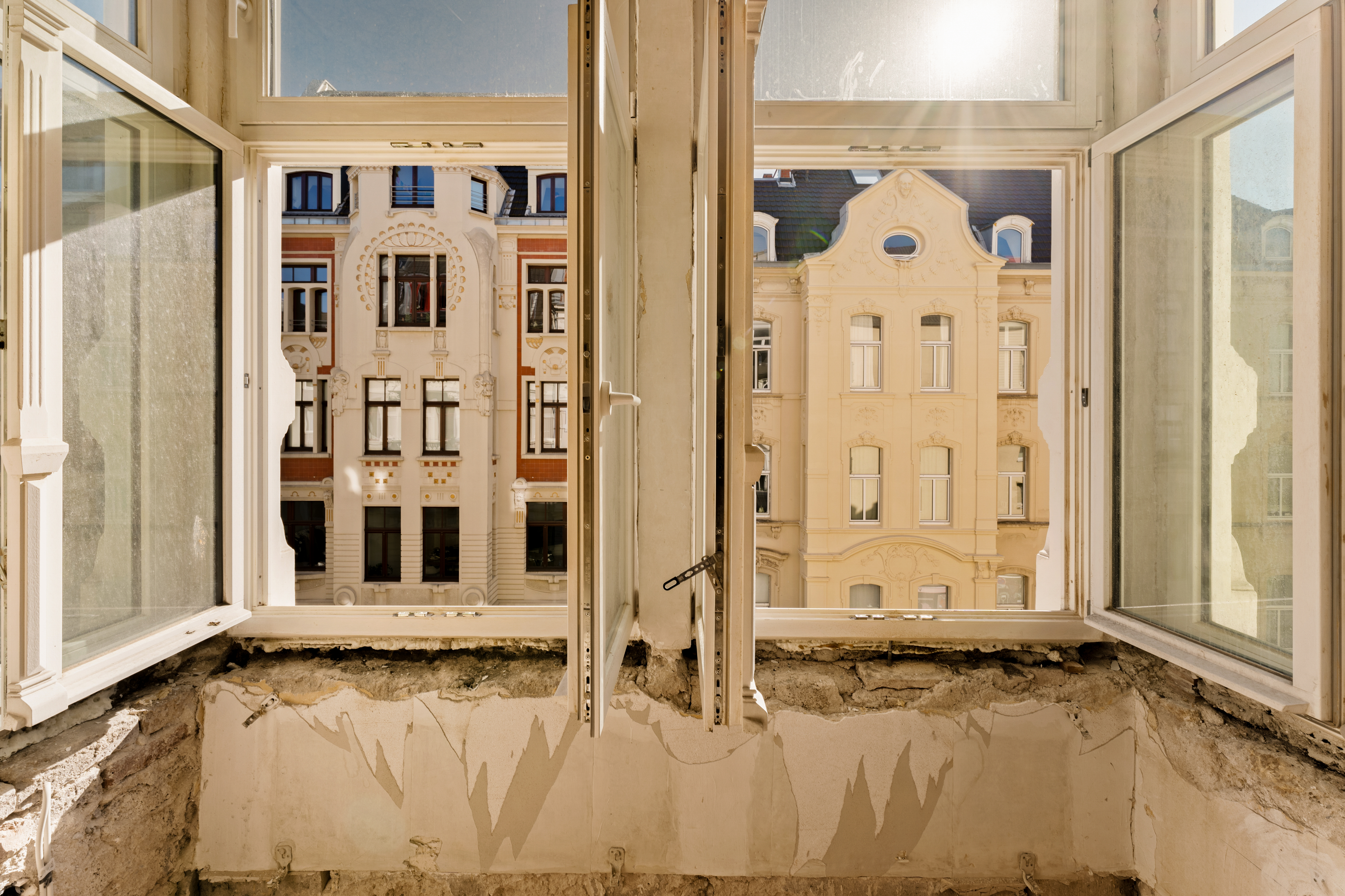 Blick aus einem renovierungsbedürftigen Raum durch offene Fenster auf Altbaufassaden.