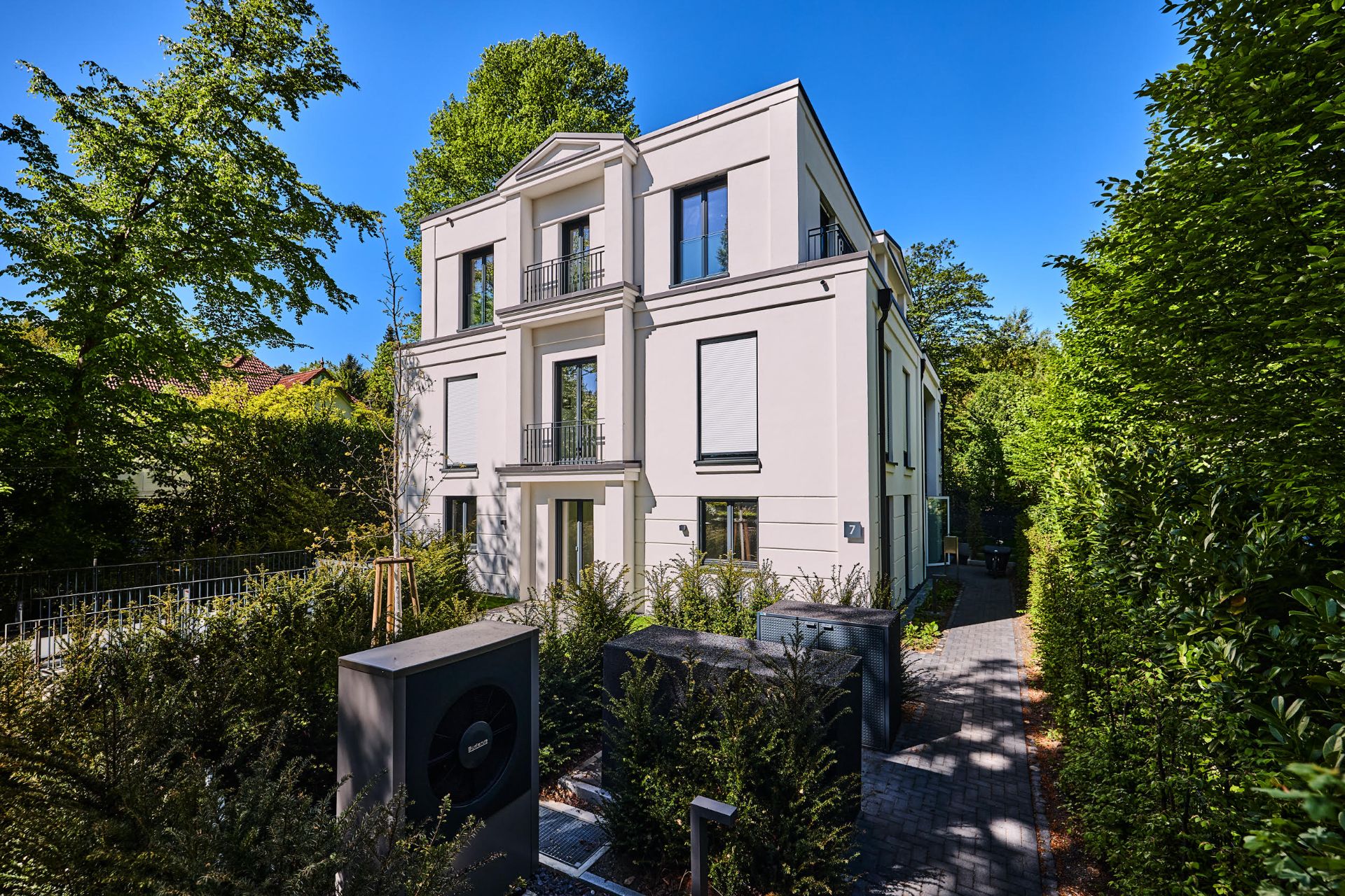 Modernes, weißes Wohnhaus mit drei Etagen, umgeben von Bäumen und einem Gartenweg.