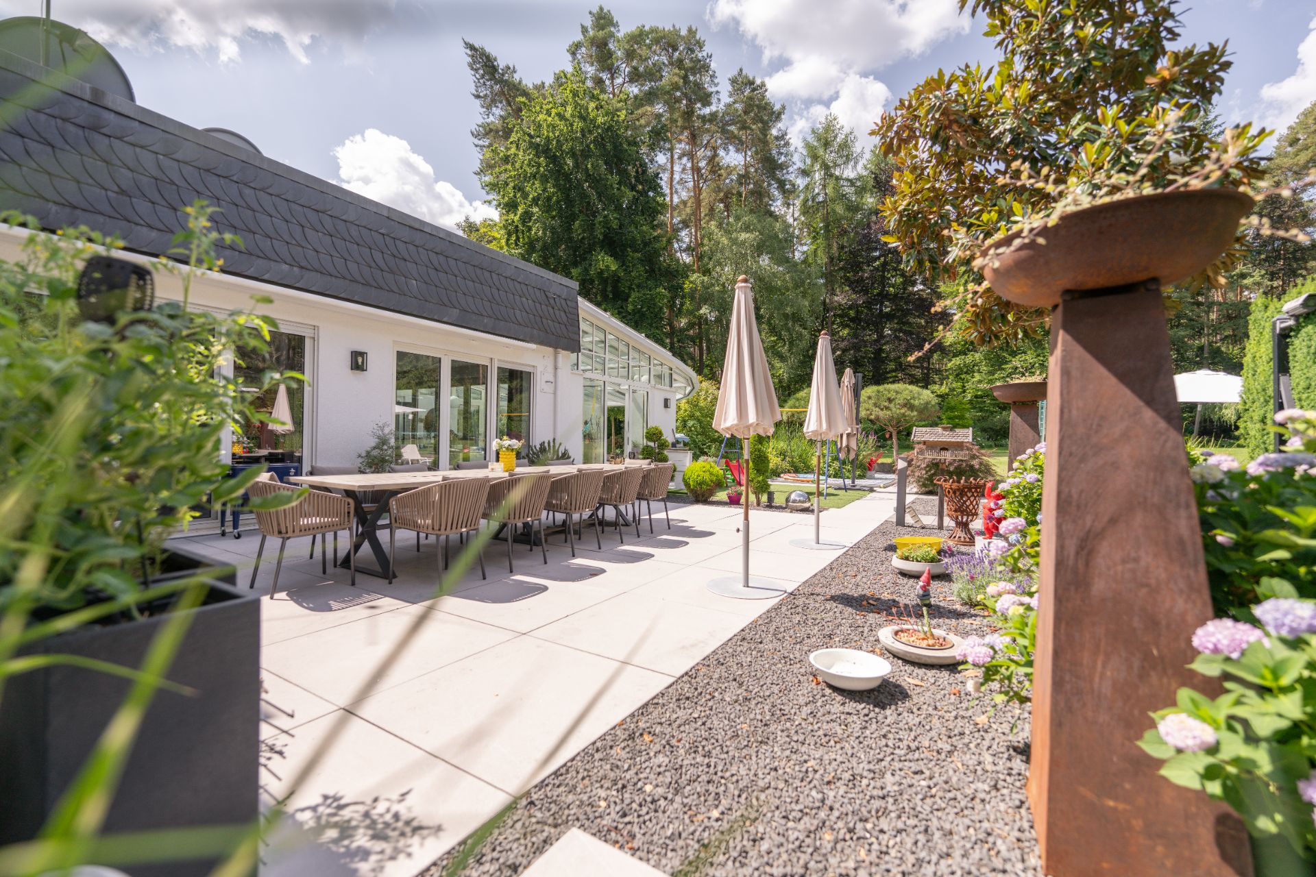 Moderne Terrasse mit Esstisch, Stühlen, Sonnenschirmen und bepflanztem Gartenbereich.