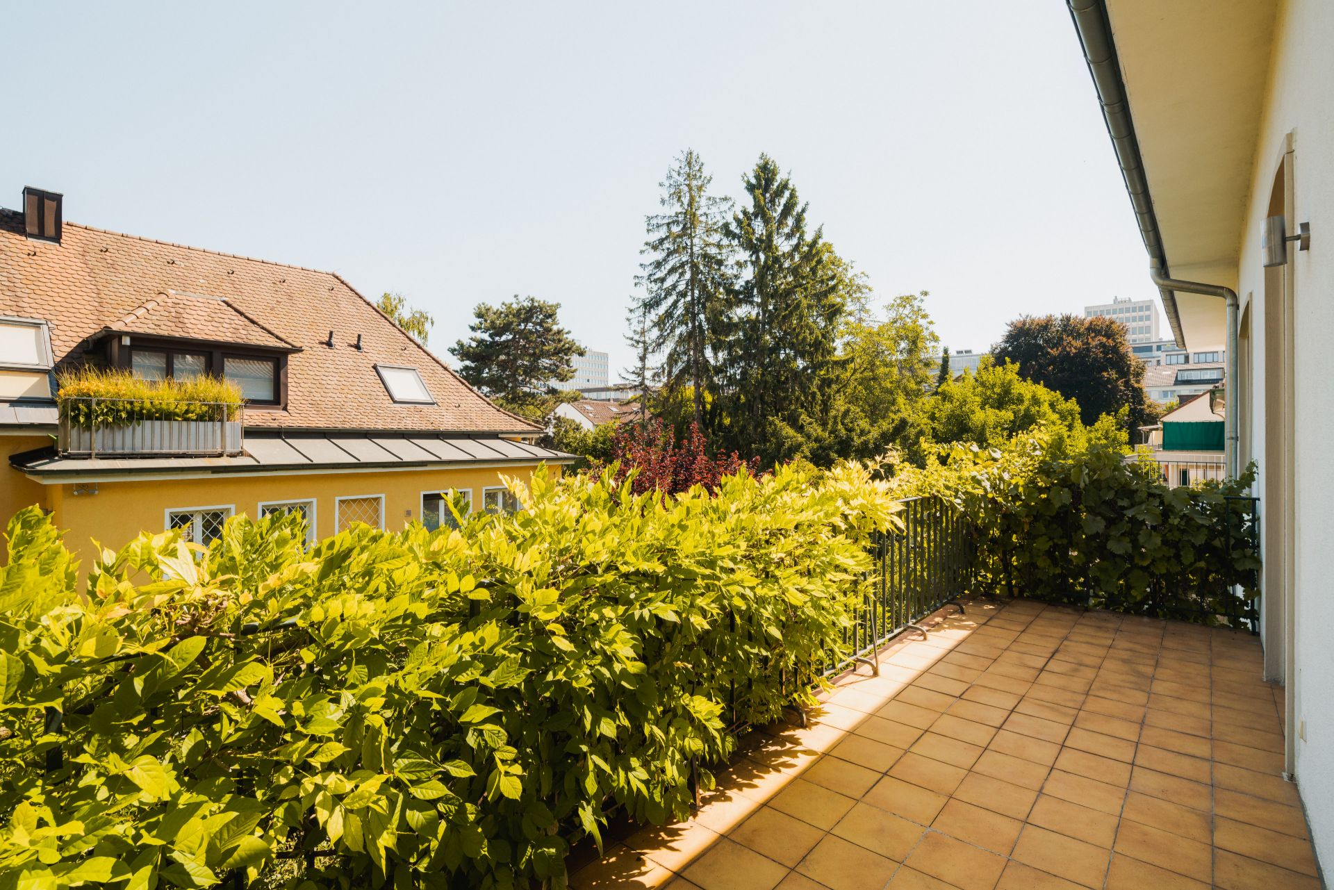 Große Terrasse mit Fliesenboden und grüner Hecke, Blick auf Nachbarhaus und Bäume.