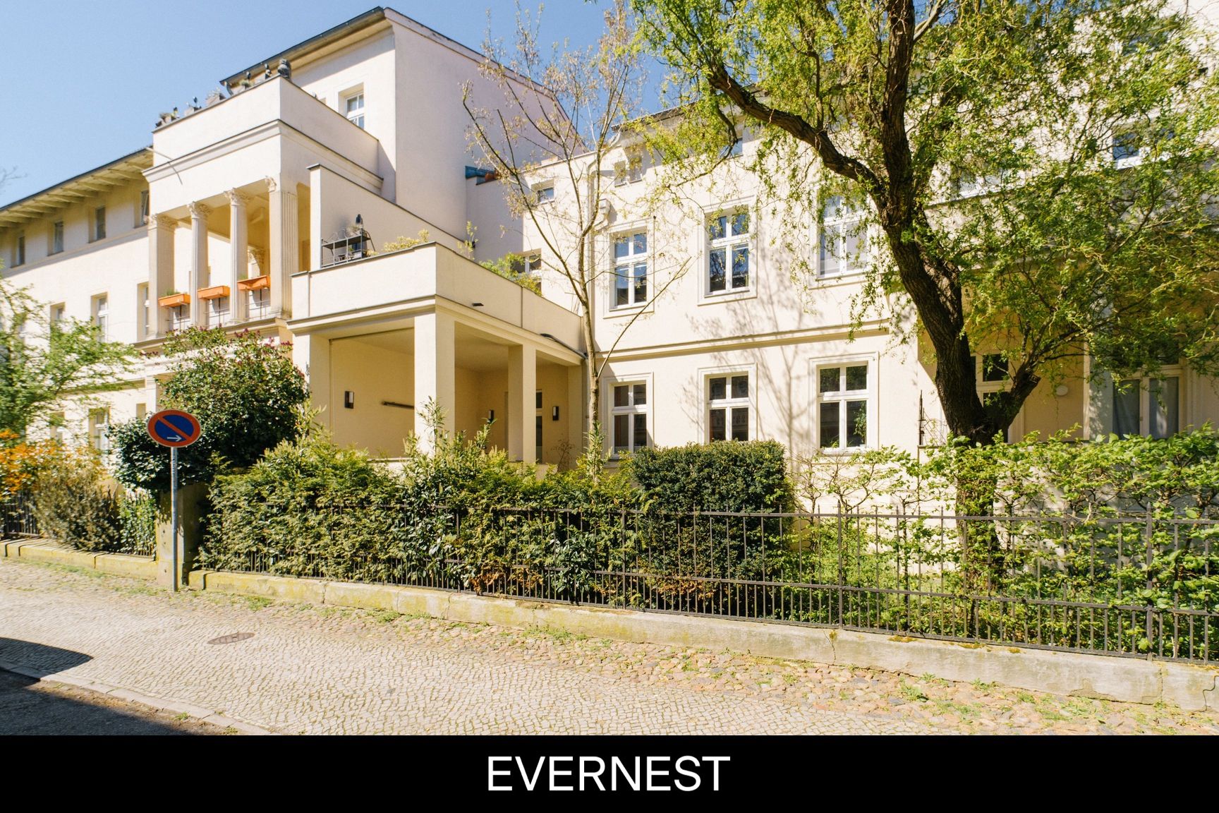 Helles Wohnhaus mit Balkon und Vorgarten, umgeben von Bäumen und Sträuchern.