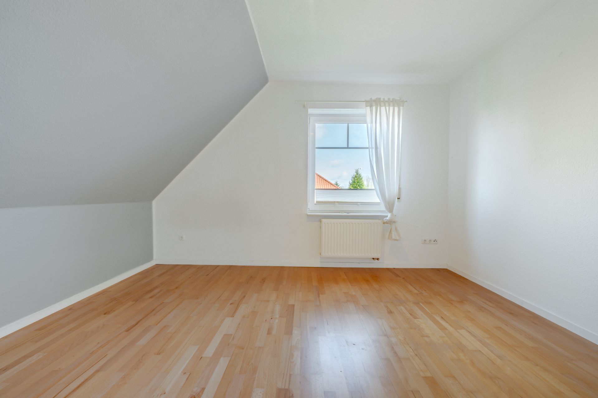 Leeres Zimmer mit schräger Decke, Holzfußboden und Fenster mit weißem Vorhang.