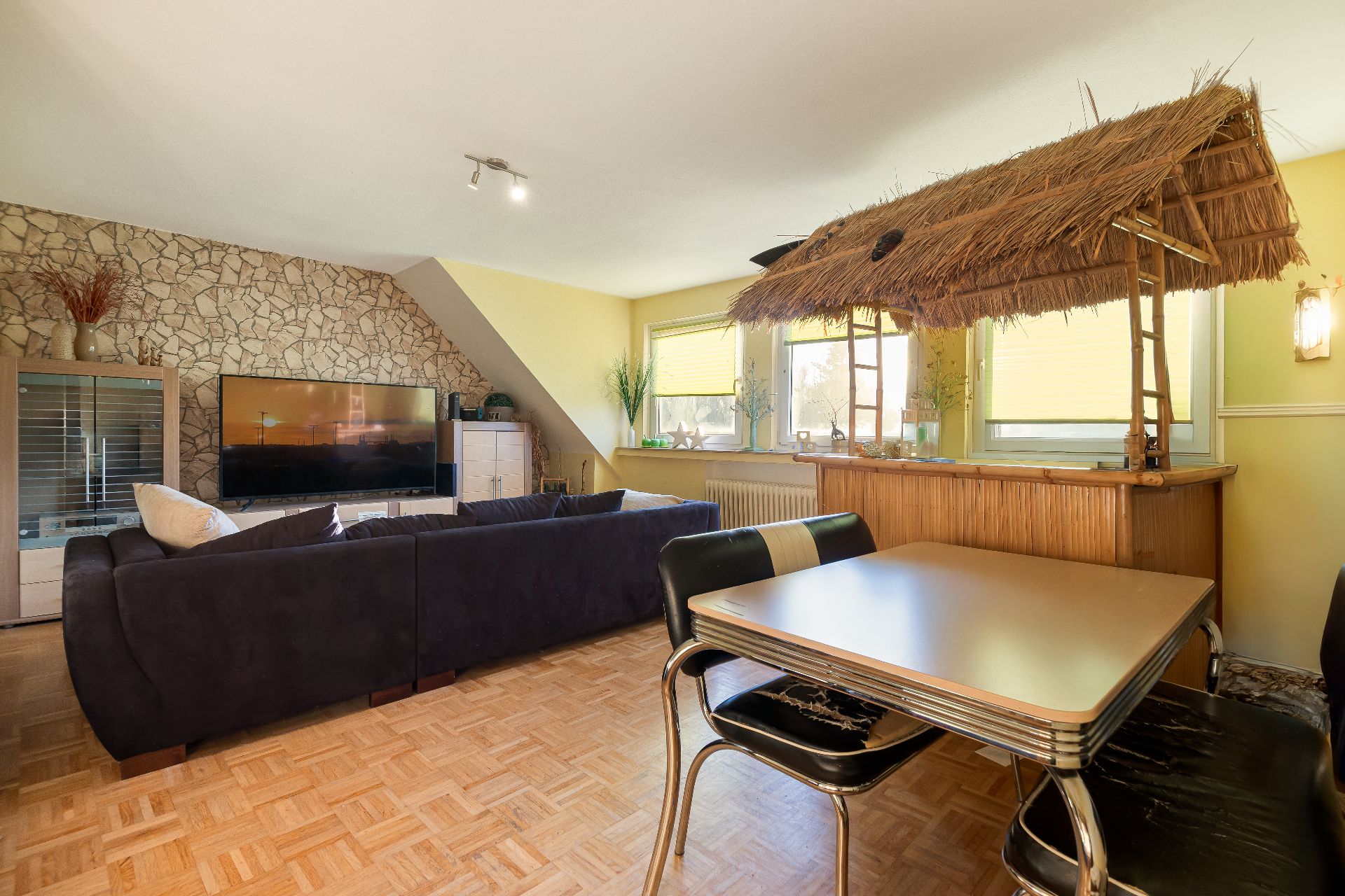 Wohnzimmer mit dunklem Sofa, Esstisch und einer Bar mit Strohdach.
