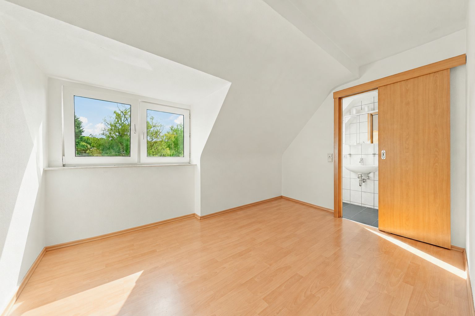 Helles Zimmer mit schräger Decke, Holzfußboden, Fenster und Blick ins kleine Bad mit Waschbecken.