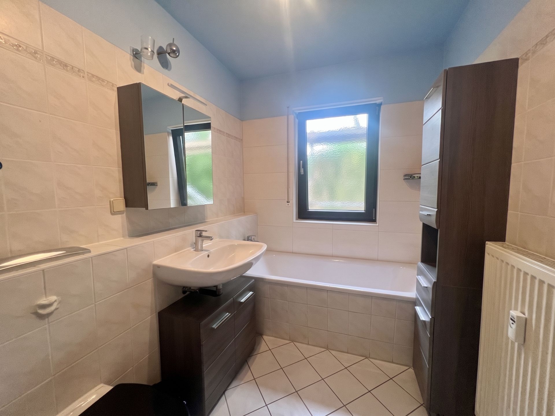 Modernes Bad mit Badewanne, Waschbecken, Spiegel, Fenster und dunklen Schränken.