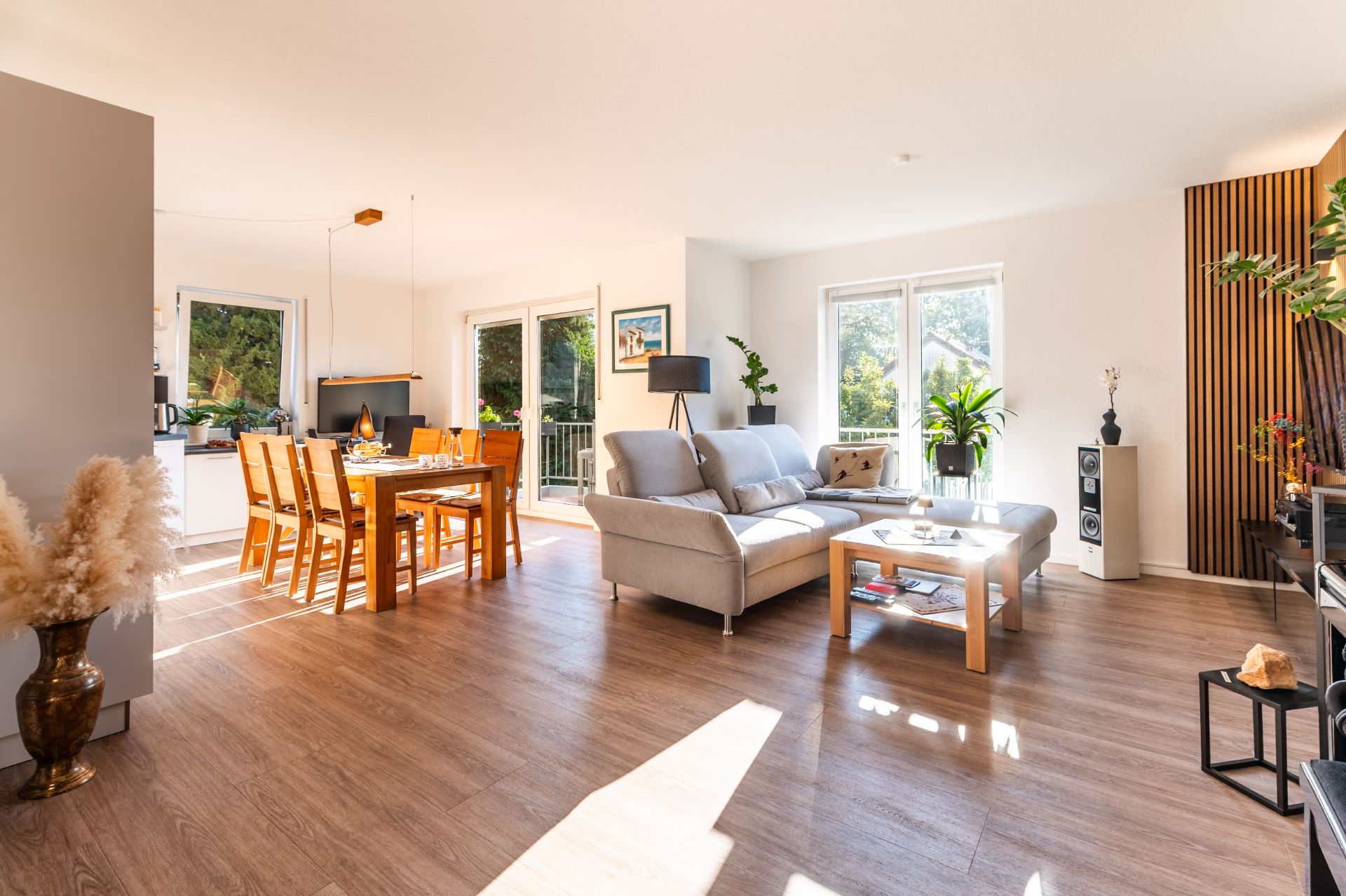 Helles Wohnzimmer mit Sofa, Esstisch, großen Fenstern und Pflanzen auf Holzfußboden.