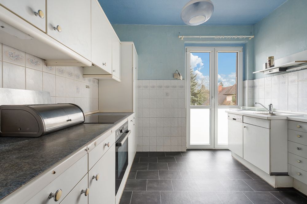 Helle Küche mit weißen Schränken, Backofen, Spüle und Fenster mit Blick nach draußen.