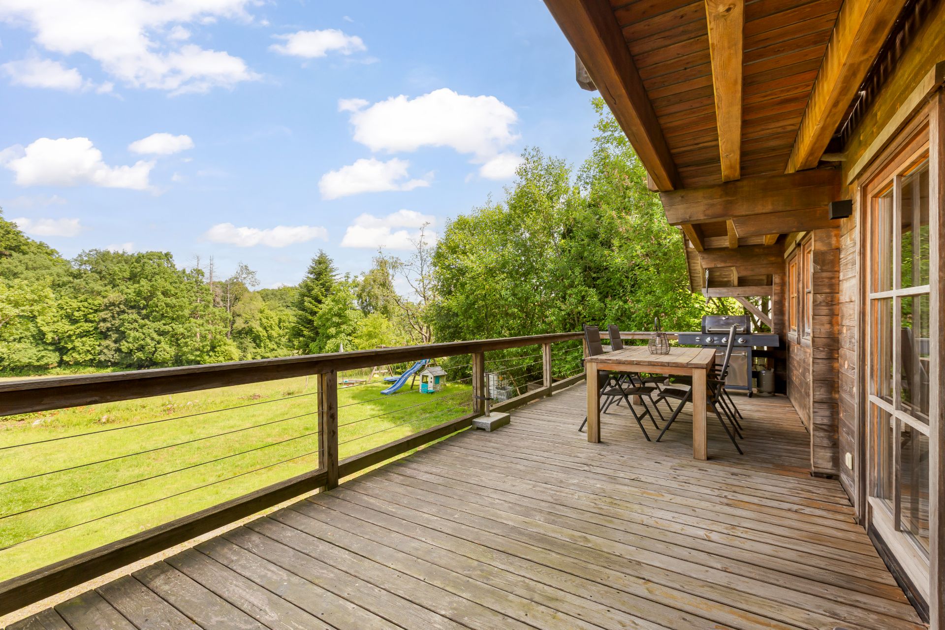 Große Holzterrasse mit Tisch und Stühlen, Blick auf grüne Wiese und Bäume.