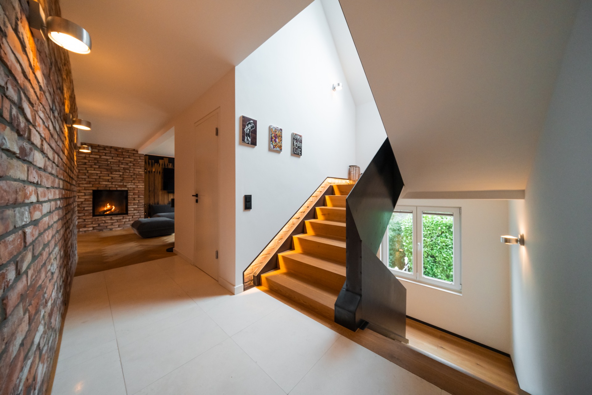 Moderne Treppe im Flur, Backsteinwand und Kamin im Wohnzimmer, großes Fenster mit Blick ins Grüne.