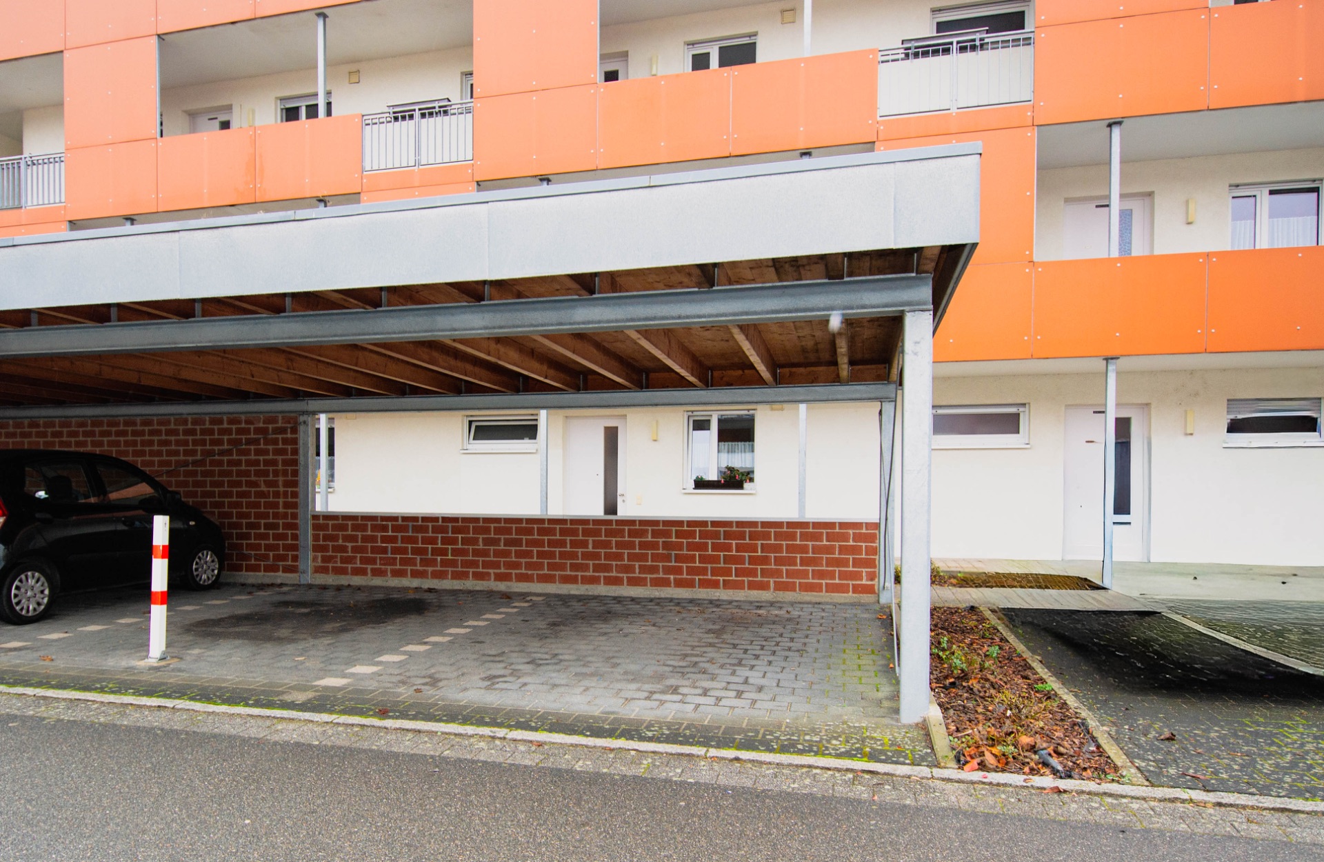 Überdachter Parkplatz mit einem Auto vor einem Wohngebäude mit orangen Balkonen.