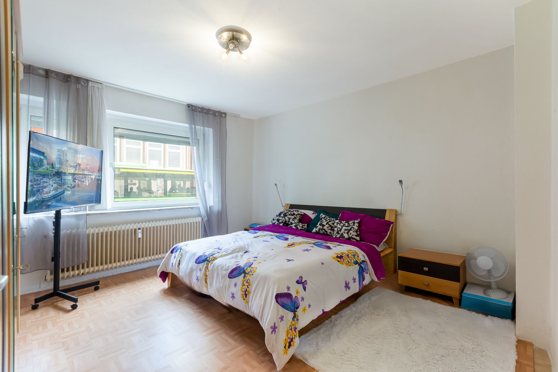 Helles Schlafzimmer mit Doppelbett, Fernseher, Nachttisch und großem Fenster mit Gardinen.