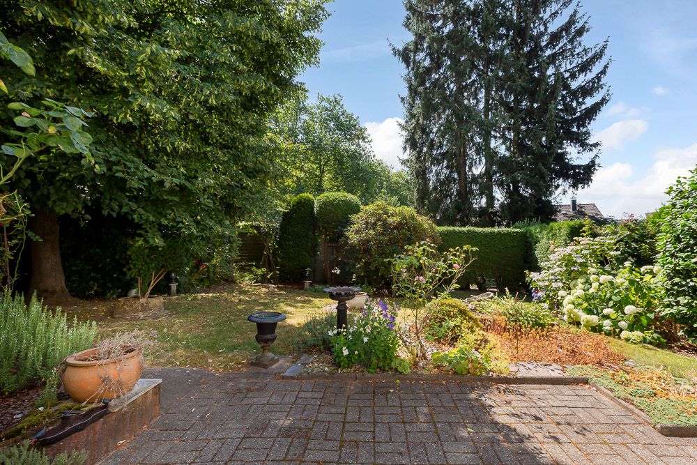 Gepflasterte Terrasse mit Blumenkübeln, Gartenbeeten und Bäumen im Hintergrund.
