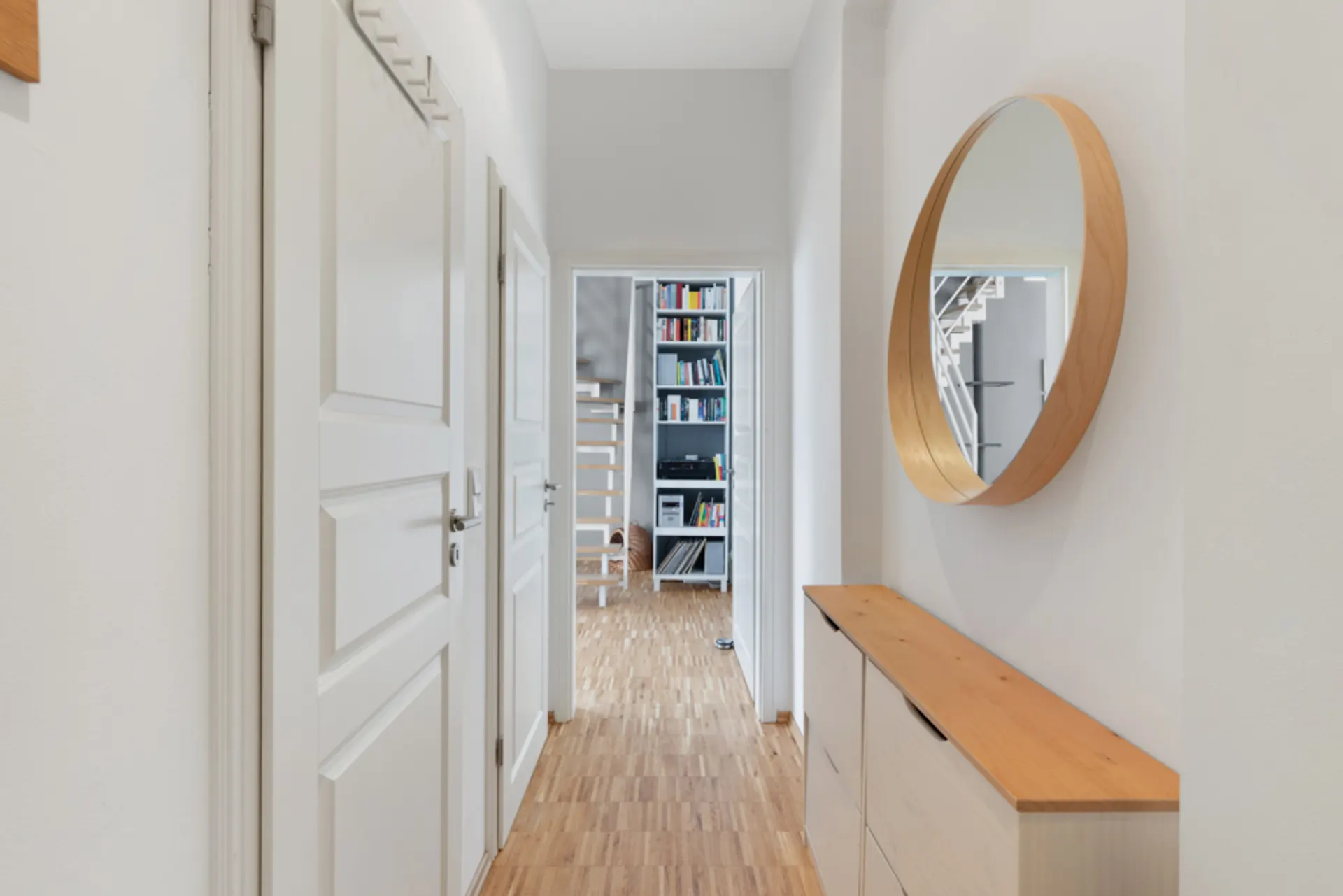 Heller Flur mit Holzboden, Kommode und rundem Spiegel. Im Hintergrund ein Regal mit Büchern.