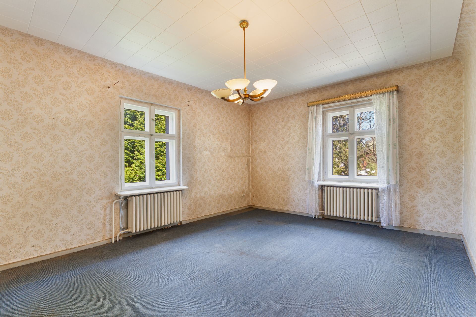Leeres Zimmer mit zwei Fenstern, Heizkörpern, hellen Wänden und einem Deckenleuchter.
