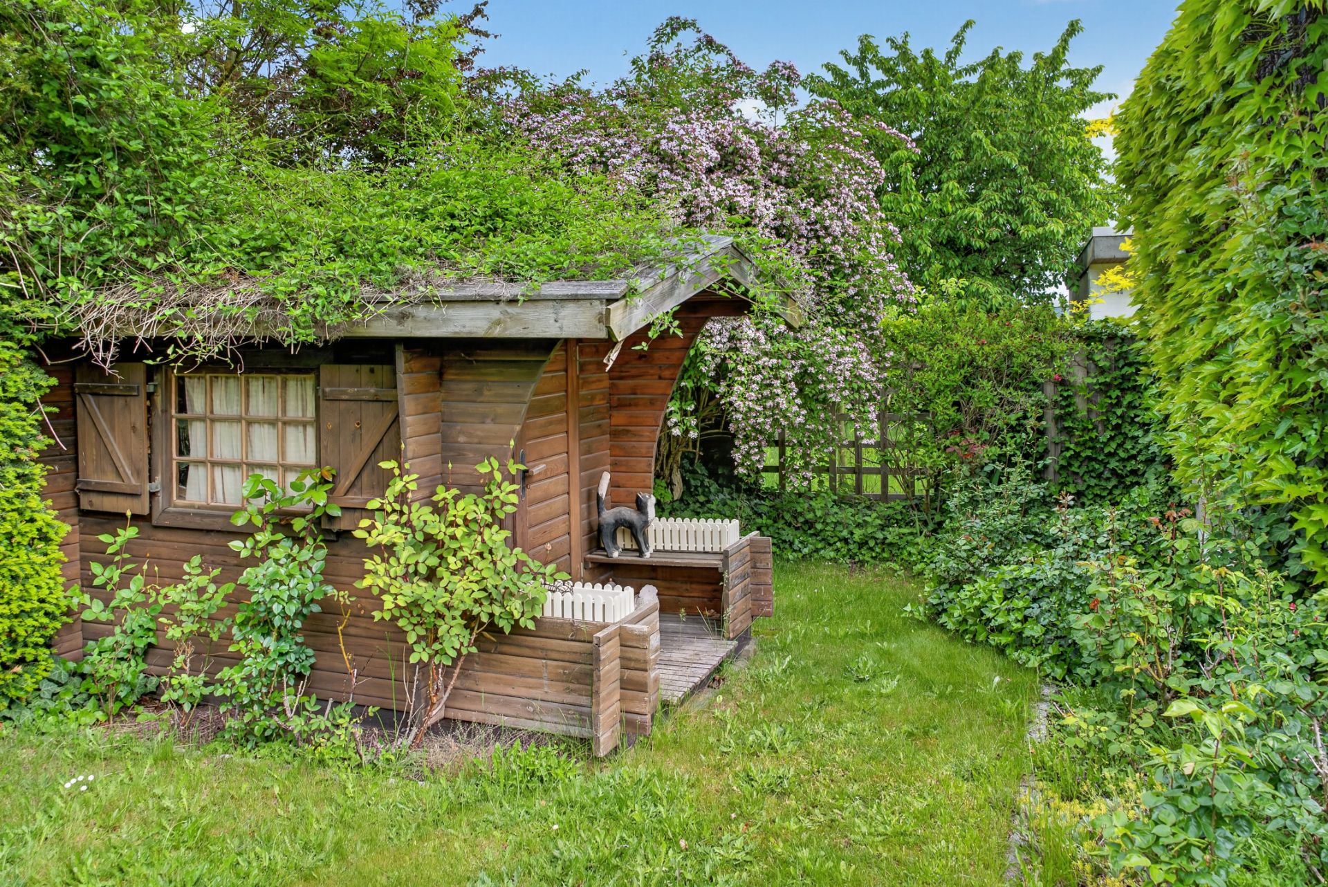 Ein kleines Holzgartenhaus umgeben von grünem Bewuchs und Rasen.