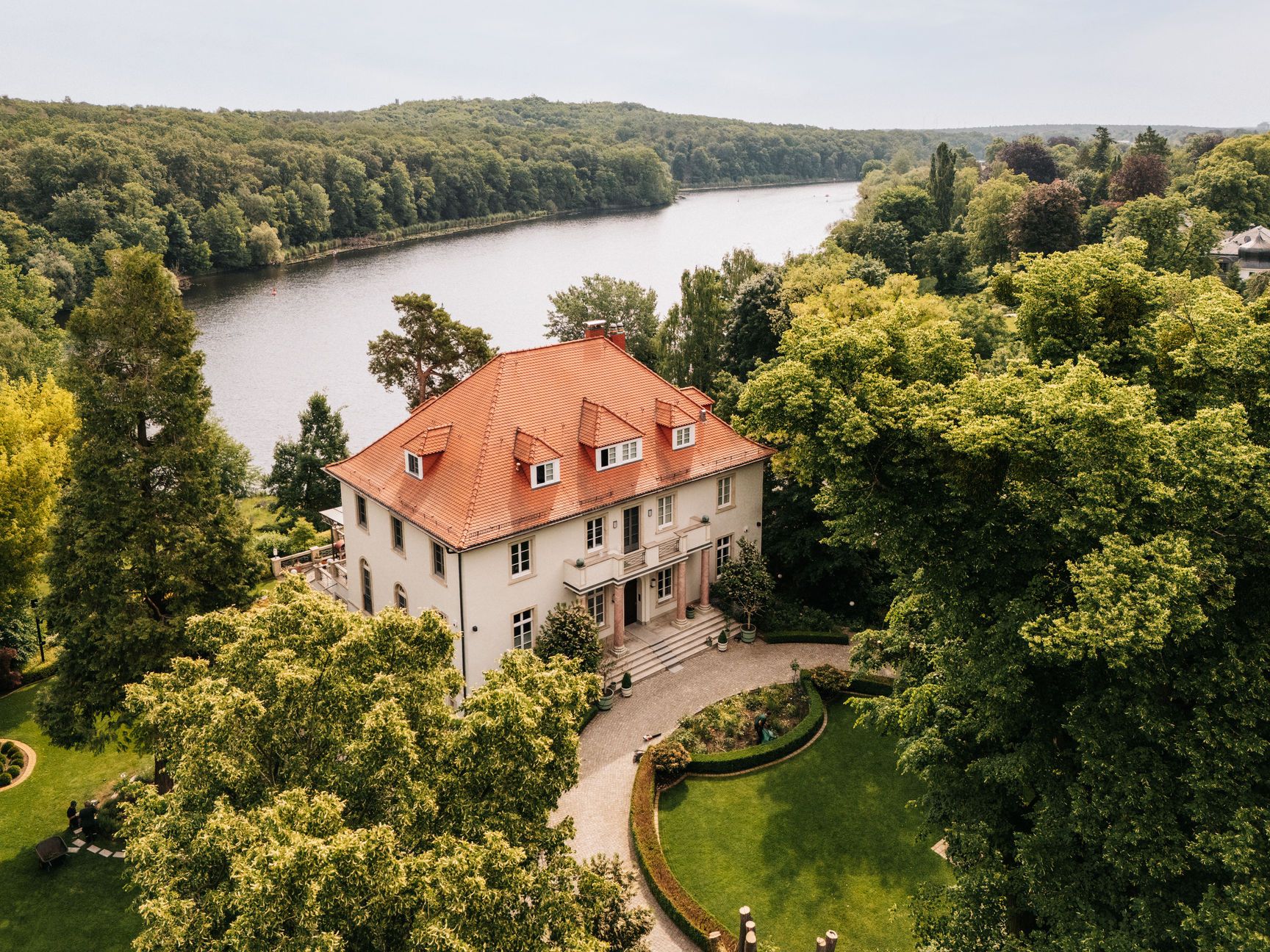 Großes Wohnhaus mit rotem Dach, umgeben von viel Grün und gelegen an einem Fluss.
