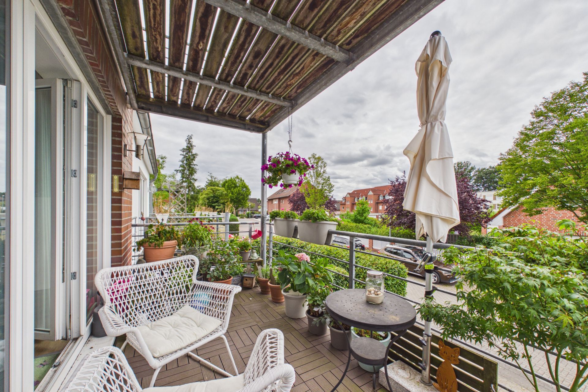 Balkon mit weißen Stühlen, Tisch, vielen Pflanzen und einem geschlossenen Sonnenschirm.
