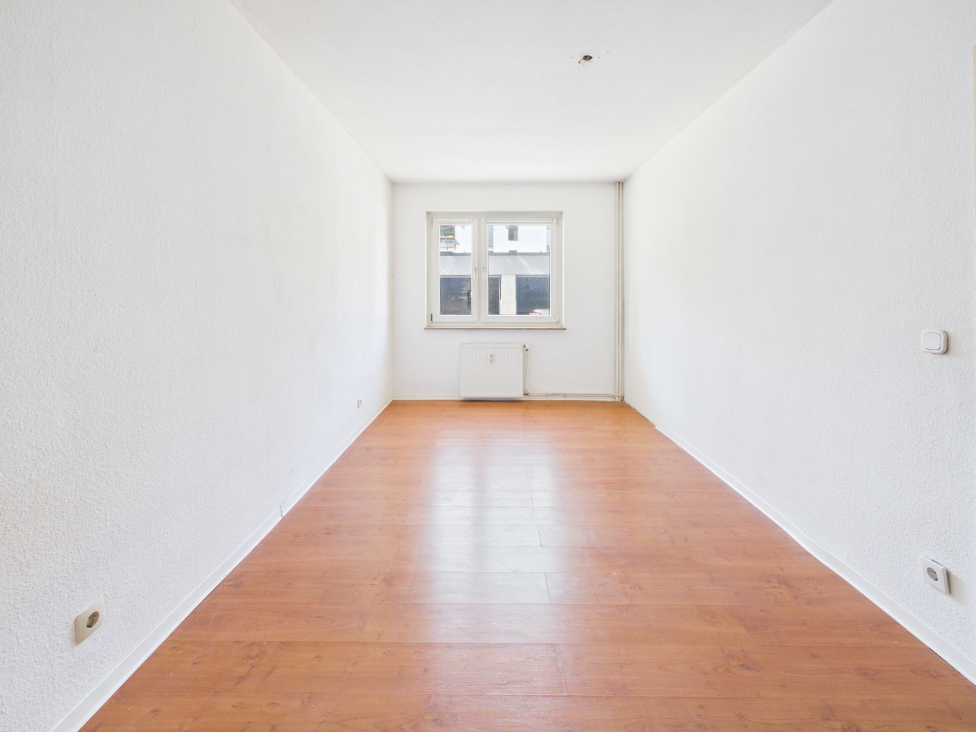 Leerer, schmaler Raum mit weißen Wänden, Holzfußboden und einem Fenster am Ende.